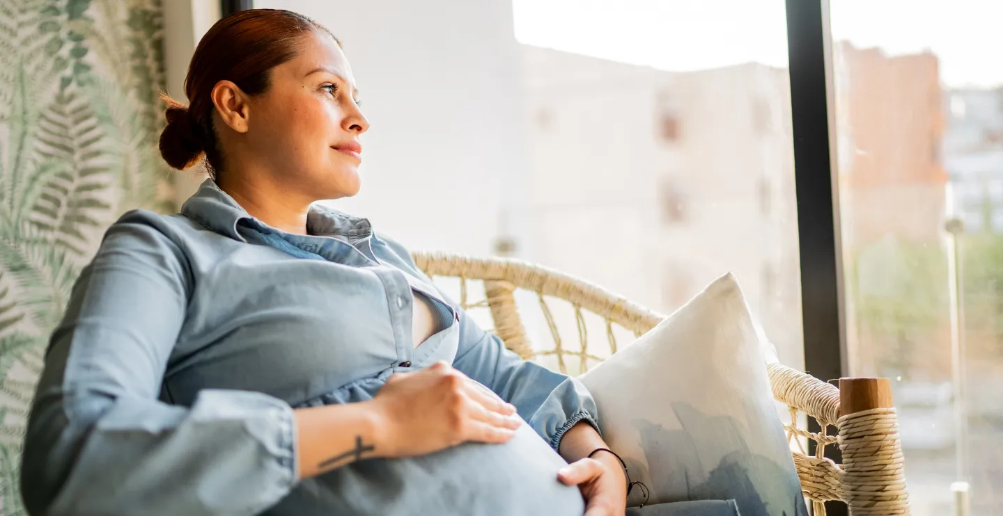 Pregnant woman relaxing in a chair by a window, gazing thoughtfully while holding her belly.