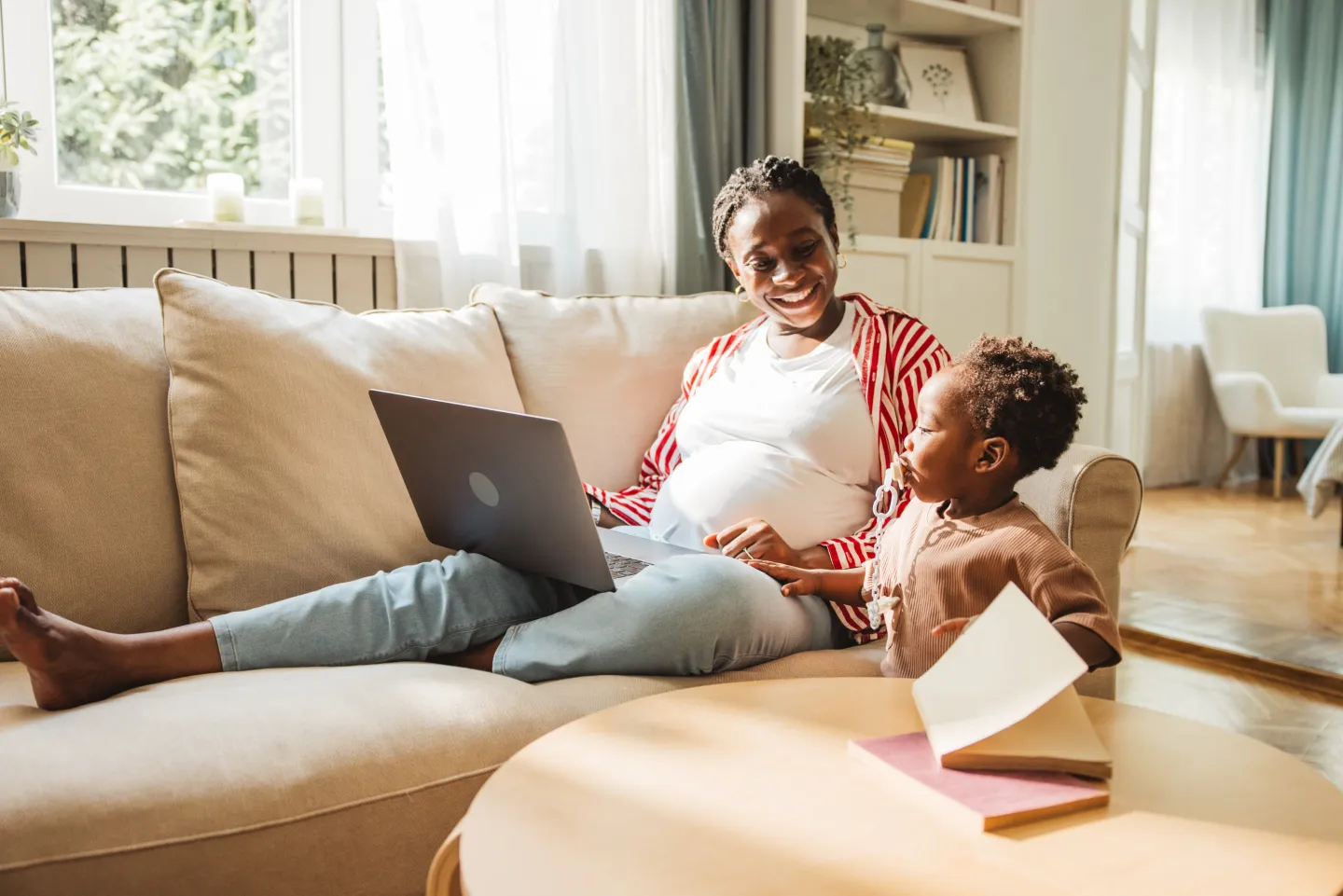 Pregnant woman smiling while using a laptop on a couch next to her young child holding a book.