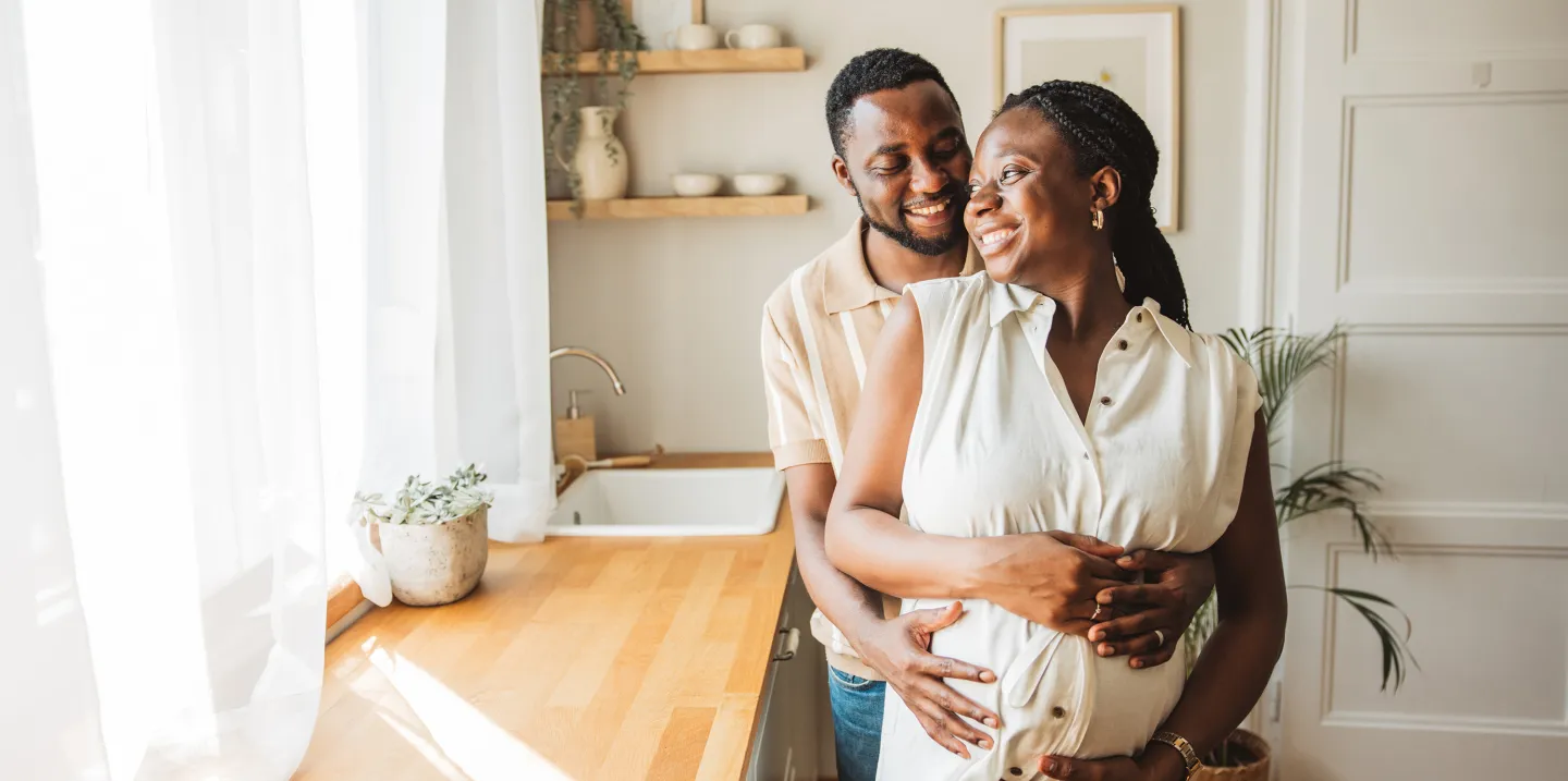 Man embracing his pregnant partner from behind as they smile together in a sunlit kitchen.