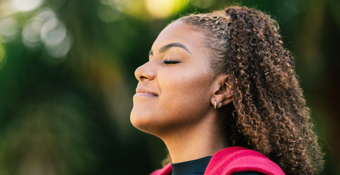 Young woman outdoors with eyes closed, smiling peacefully as she takes a deep breath.
