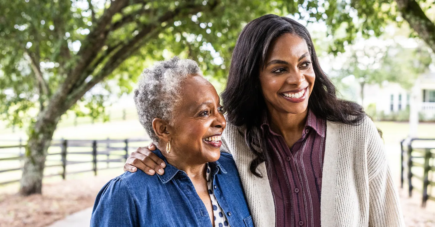Smiling older woman walking arm-in-arm with a younger woman outdoors beneath leafy trees.