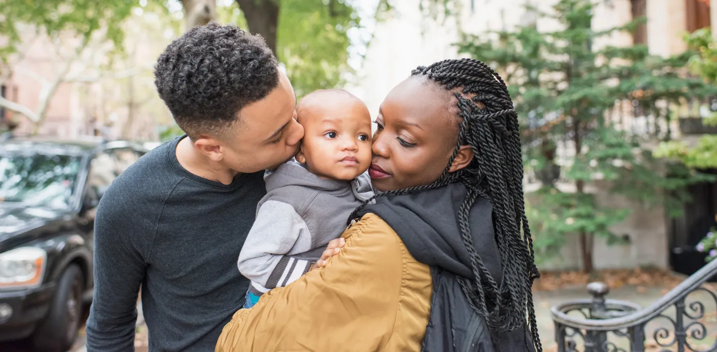 A young couple lovingly embraces their baby on a tree-lined city street, with the father kissing the child's cheek and the mother holding them close.