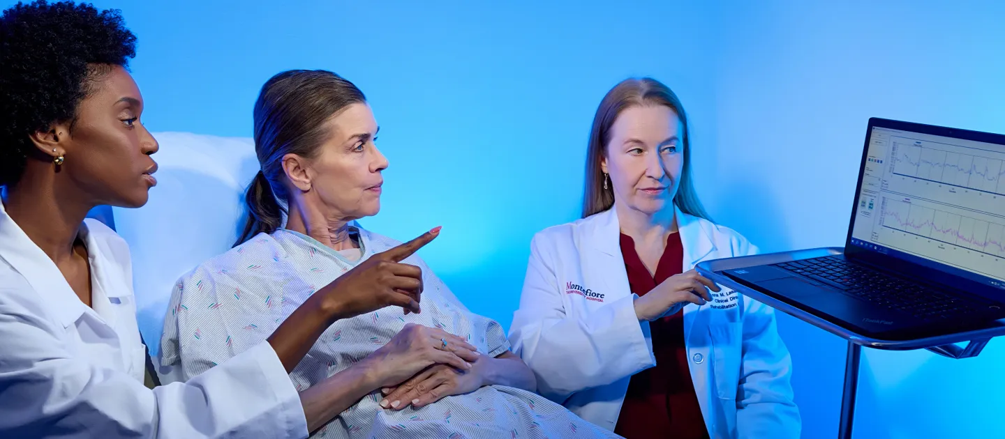 Female doctors review patient data on a laptop with a woman in a hospital gown during a rehabilitation consultation.