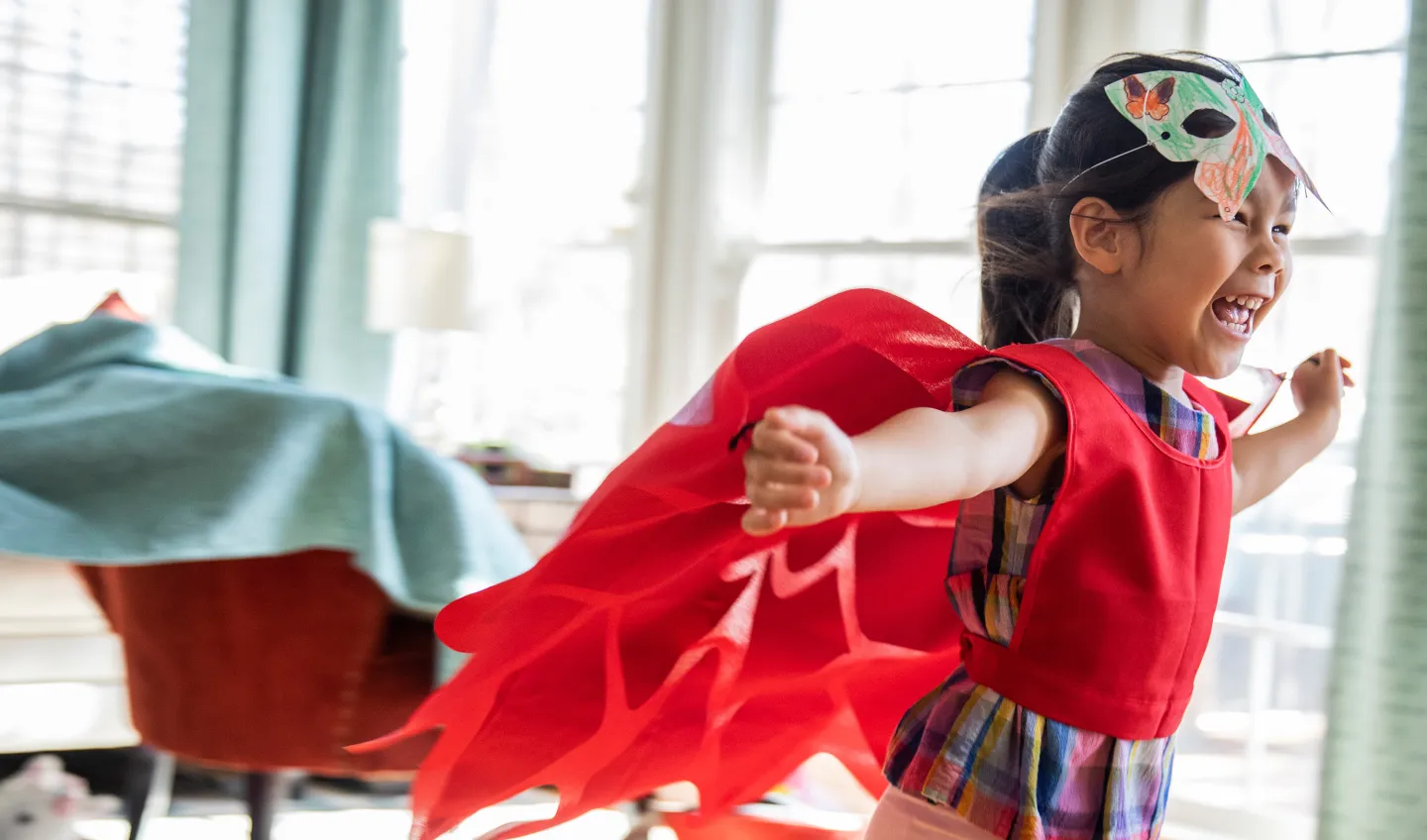 Young girl smiling and running indoors wearing a red cape and handmade butterfly mask.