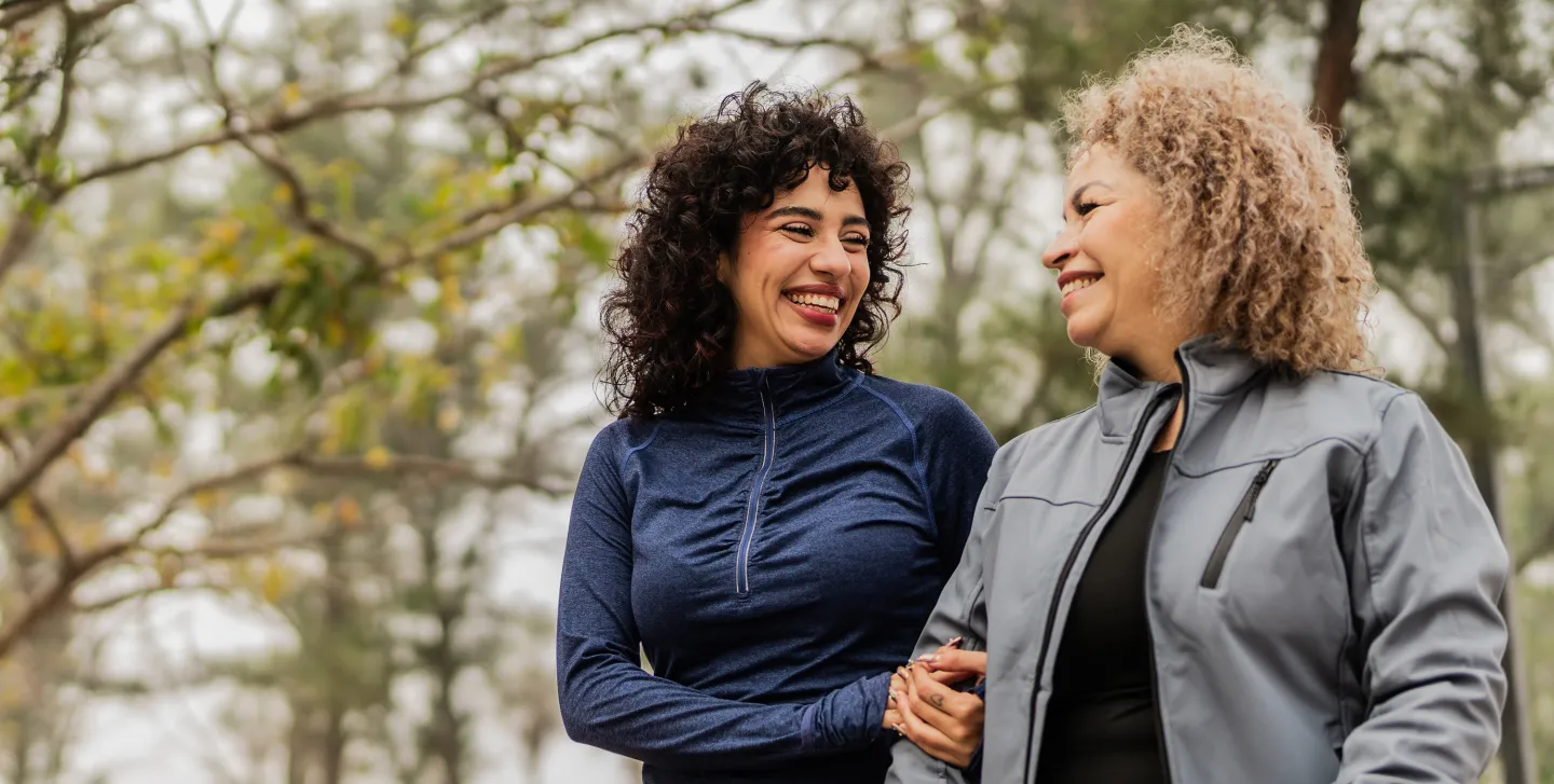 Two women walking together outside, smiling and holding each other's arms in a friendly gesture.