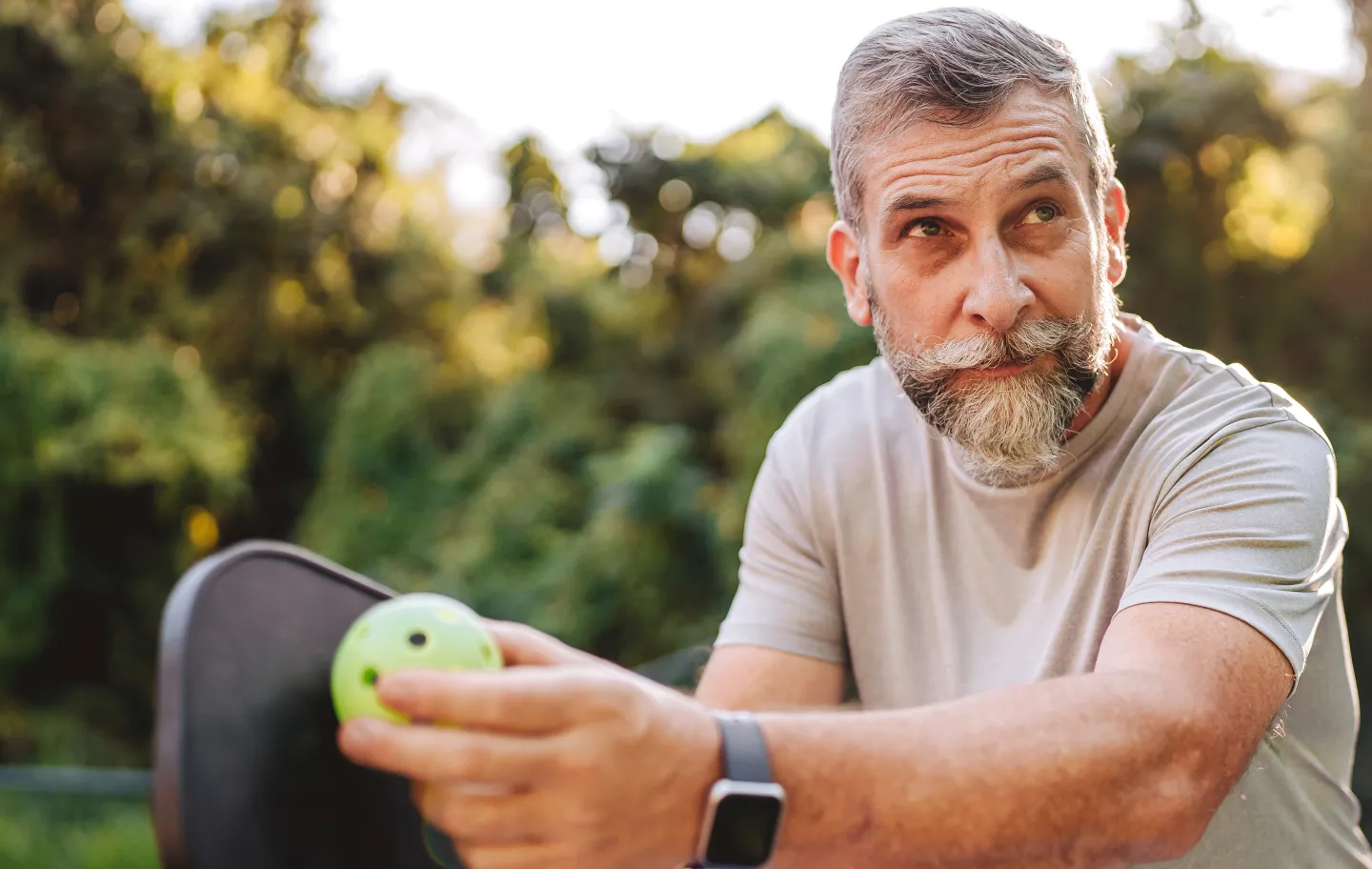 Older man outdoors holding a pickleball paddle and ball, preparing to play with a focused expression.