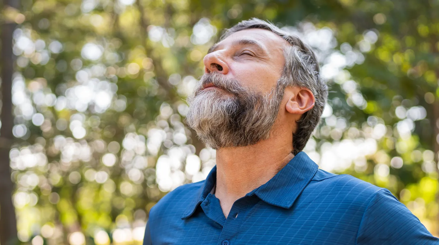 Bearded man outdoors with eyes closed, taking a deep breath in a peaceful forest setting.