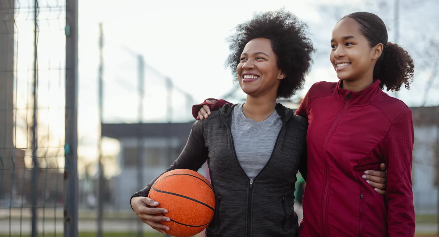 A smiling woman with her arm around a young girl holds a basketball on an outdoor court, symbolizing family, sports, and togetherness.