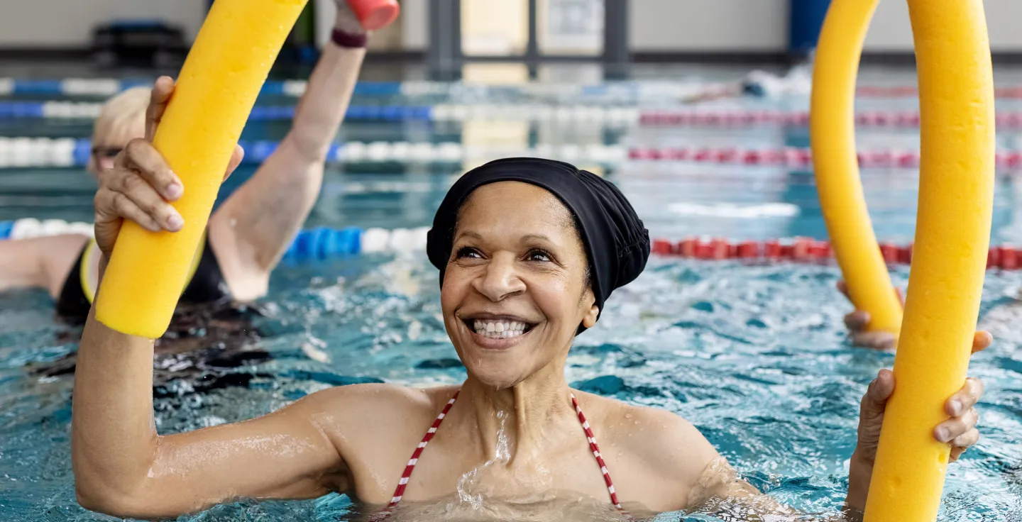 Smiling woman in a pool holding yellow foam noodles during a water exercise class.