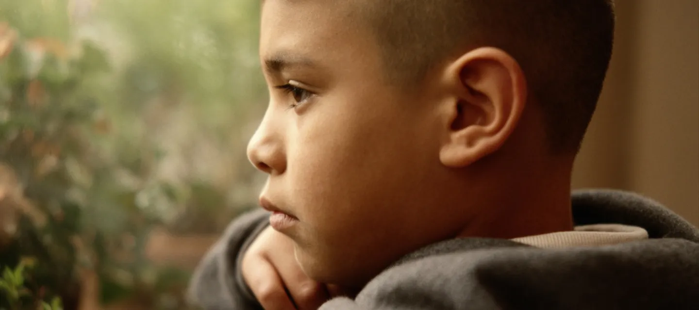 Young boy in gray hooded sweatshirt indoors looking out a window.