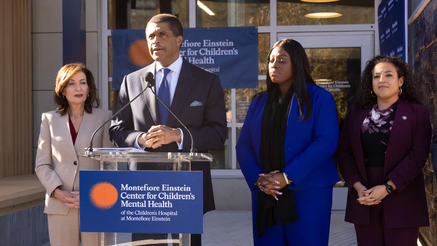 Dr. Philip Ozuah speaks at a podium bearing the Montefiore Einstein Center for Children's Mental Health logo, flanked by New York Governor Kathy Hochul on his right and Bronx Borough President Vanessa Gibson and State Senator Nathalia Fernandez on his left, outside the center's entrance.