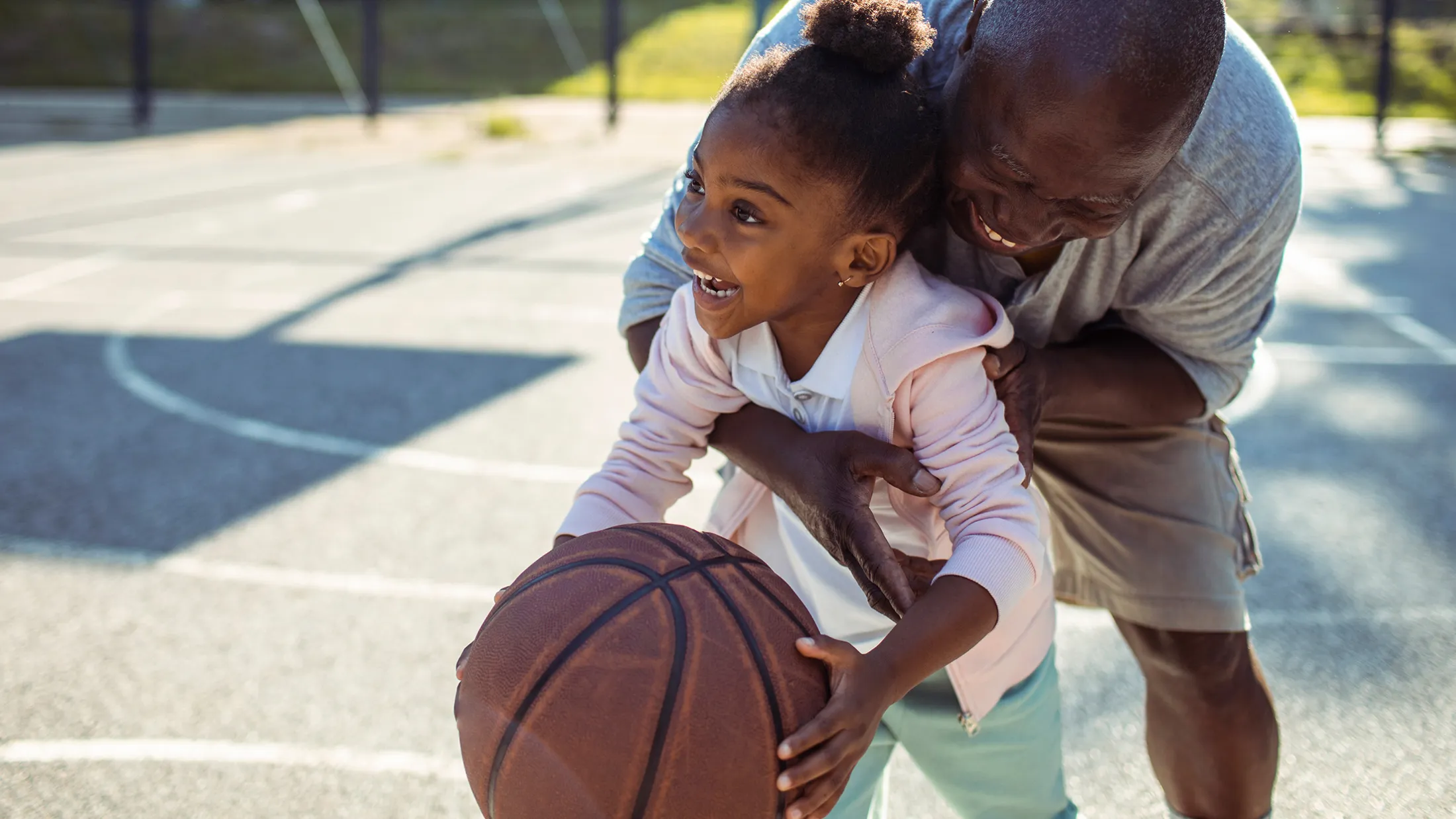 A grandfather and granddaughter playing basketball together on an outdoors basketball court.