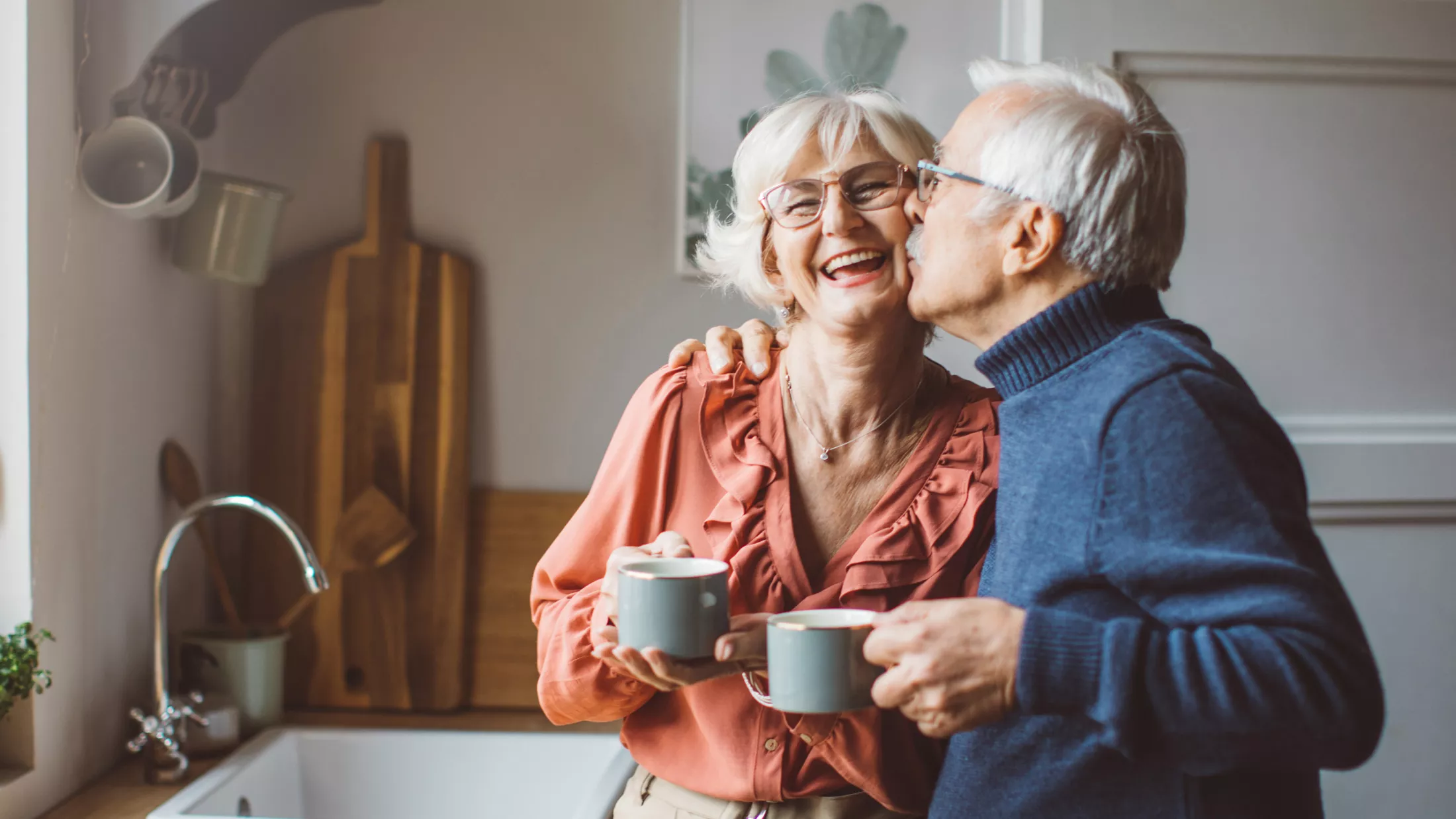 A senior couple standing in front of the kitchen window and drinking tea.
