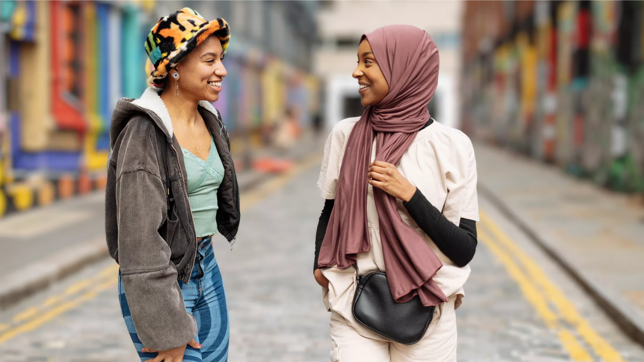 Two young women laugh together on a brightly decorated city street.