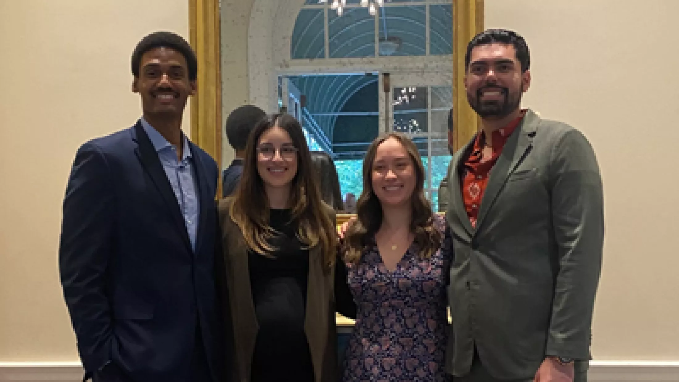 Four people posing indoors in front of a mirror, dressed in business or semi-formal clothing.