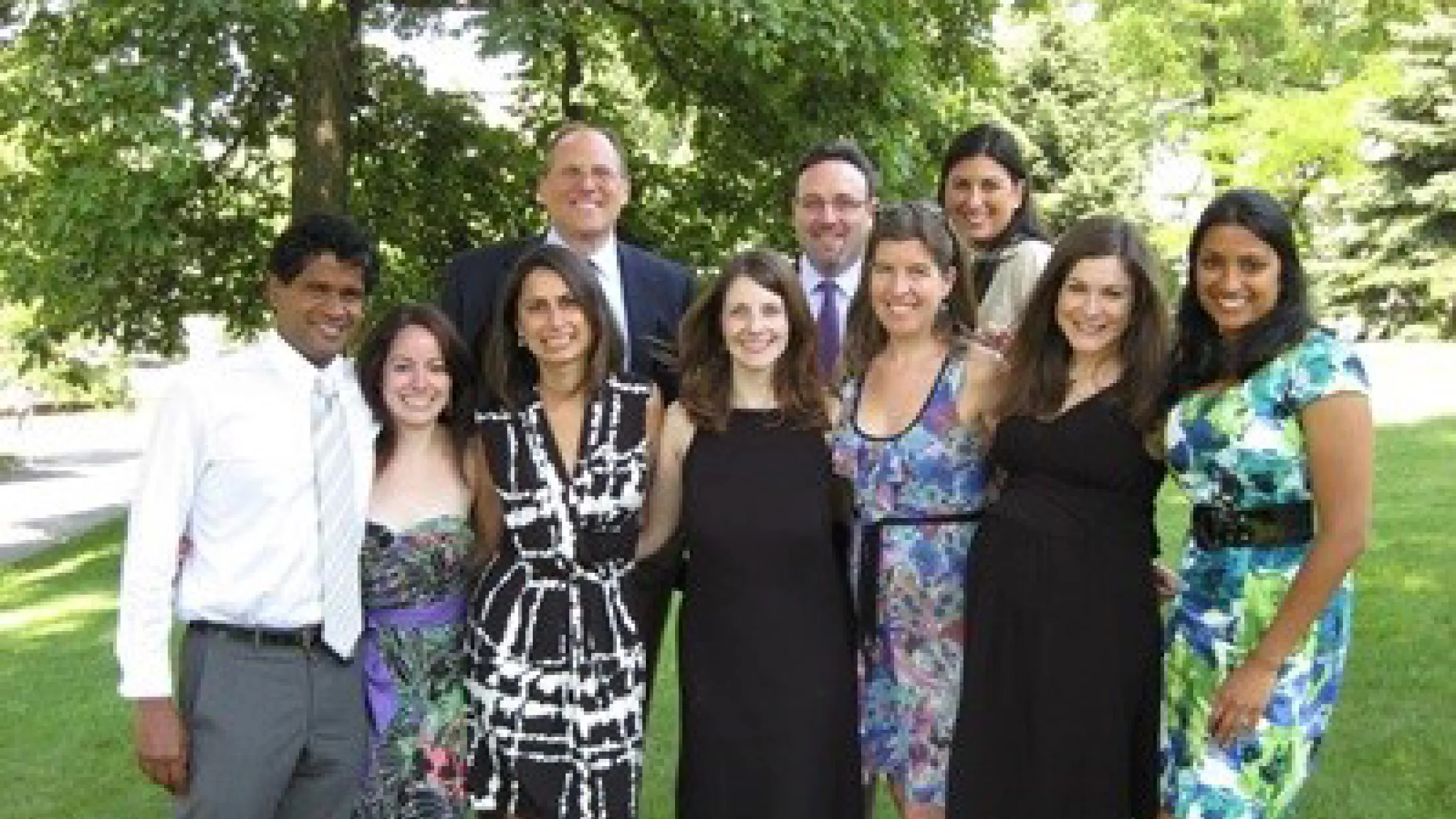 Group of people dressed formally standing on grass under trees in sunny weather.