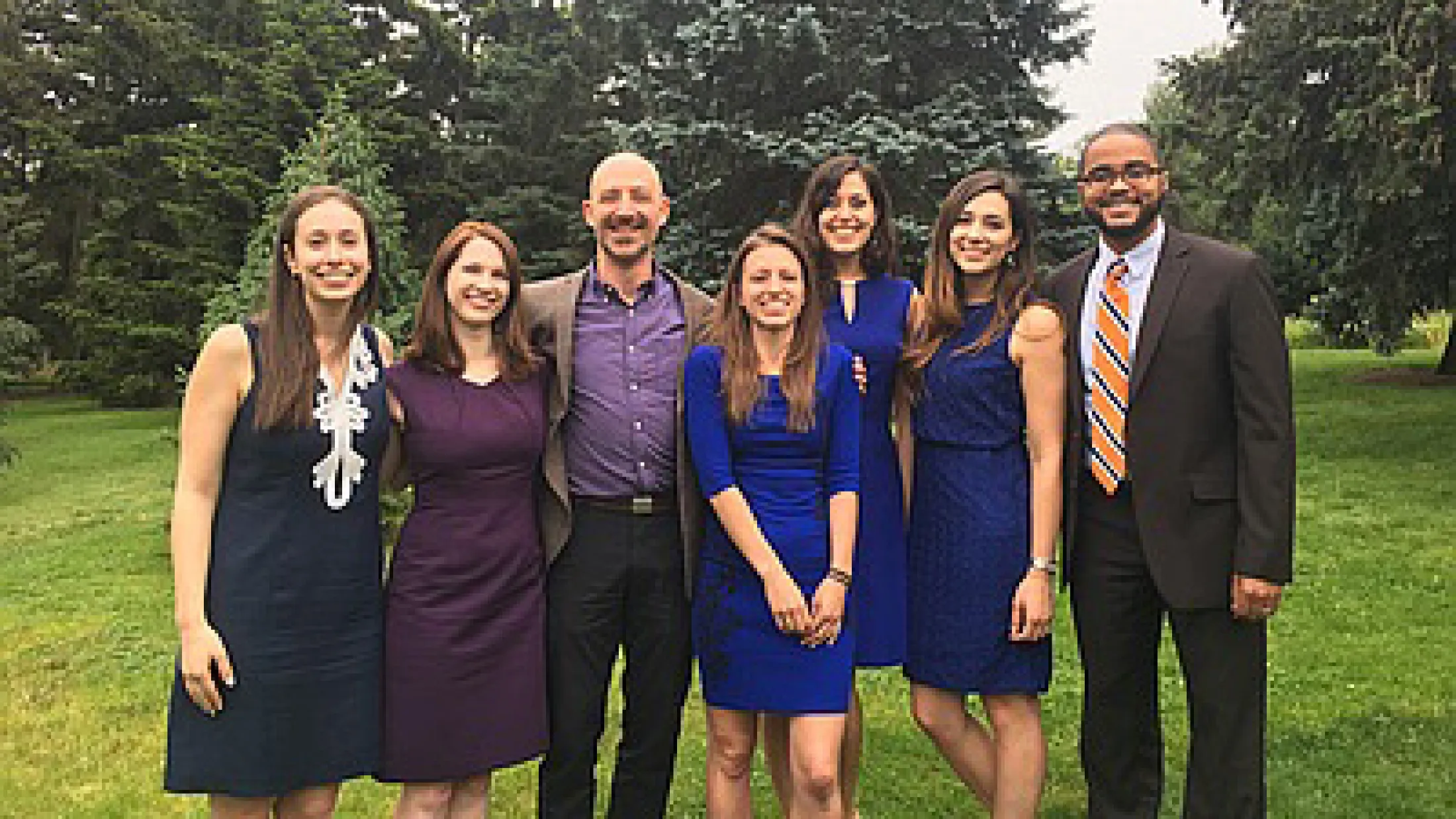 Seven people smiling outdoors in formal attire, standing on grass with trees behind them.