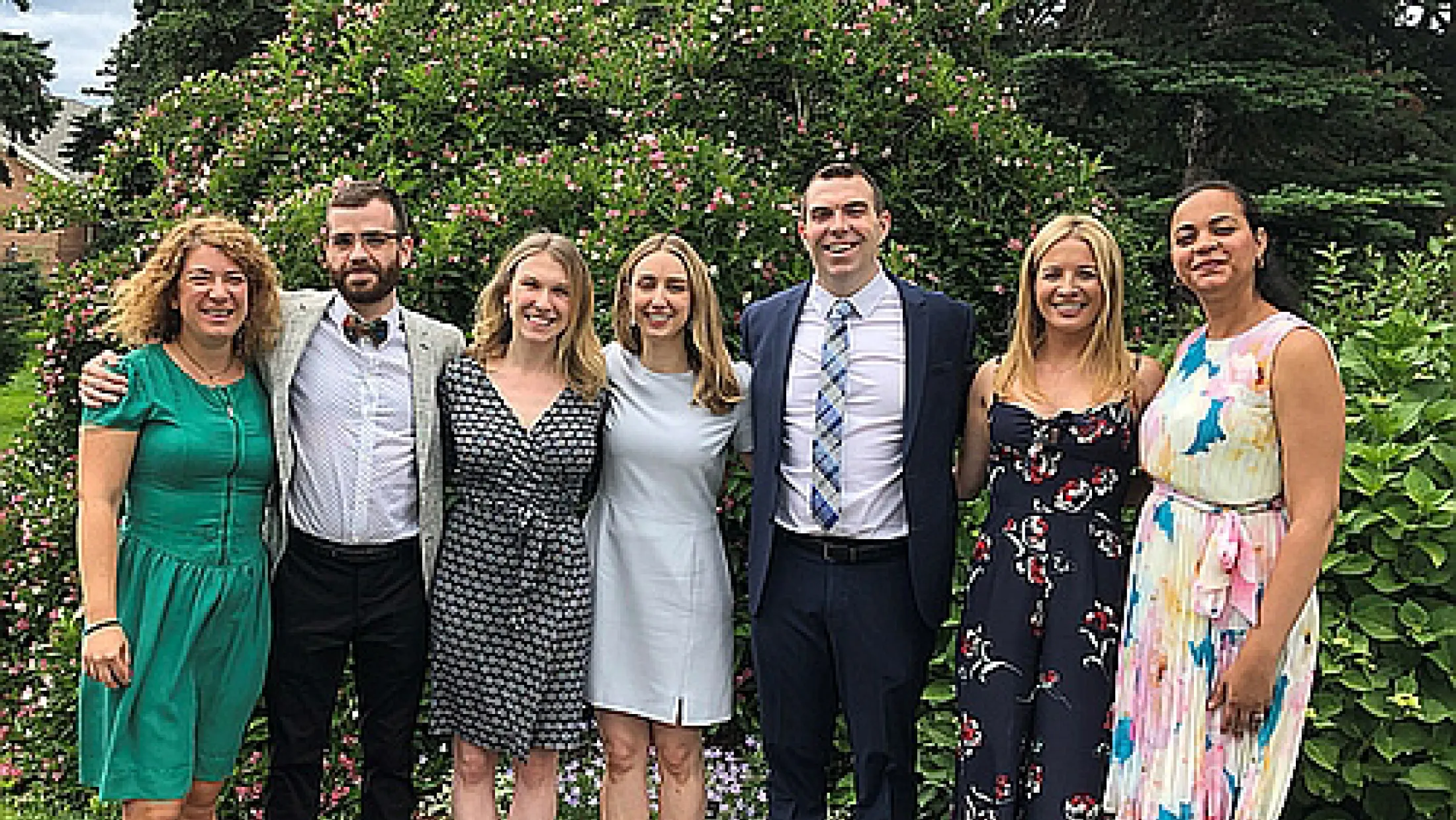 Group of seven people smiling and dressed up in colorful attire with greenery behind them.