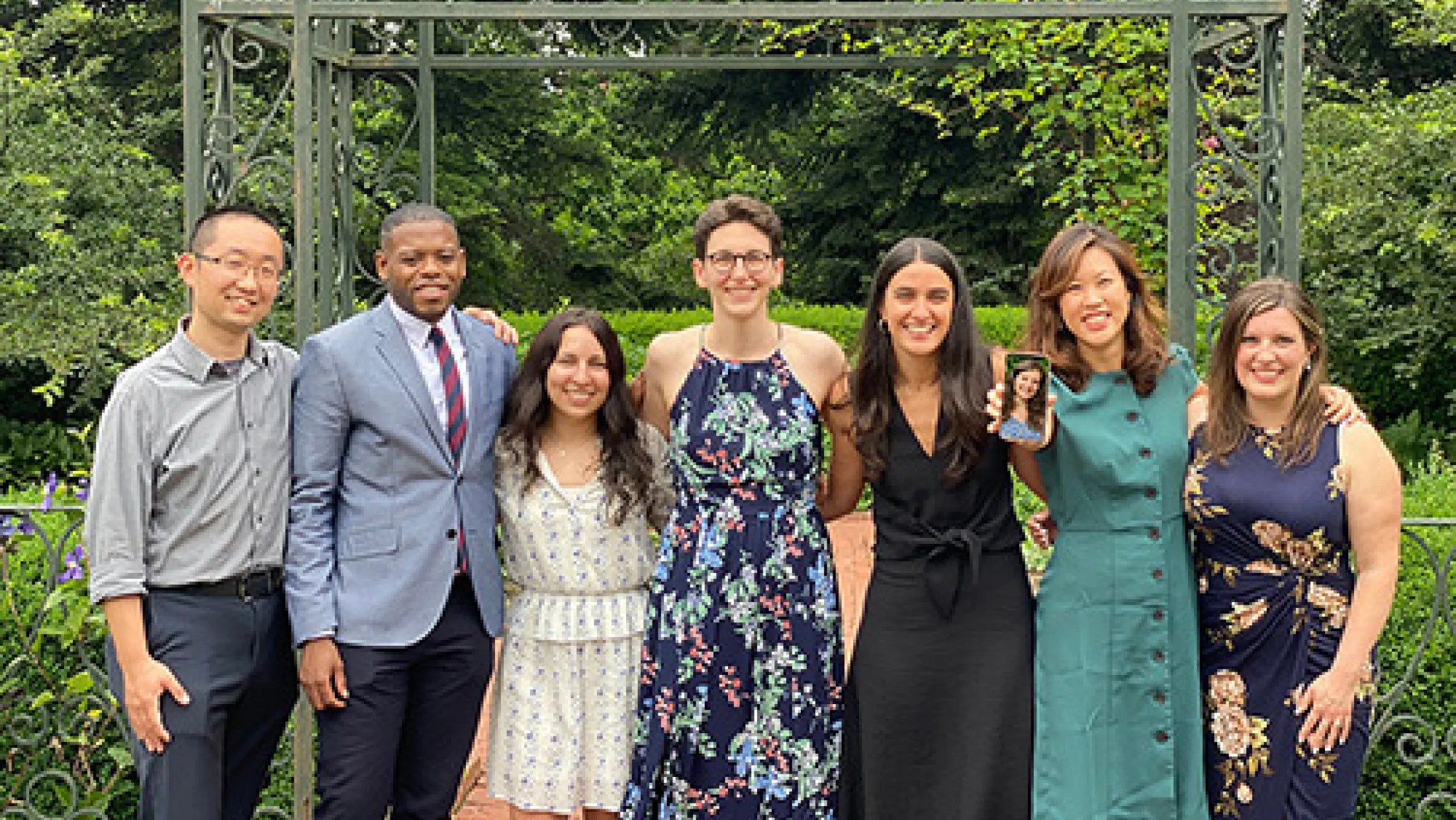 Seven people posing outdoors in front of a metal gazebo structure, dressed in formal wear.
