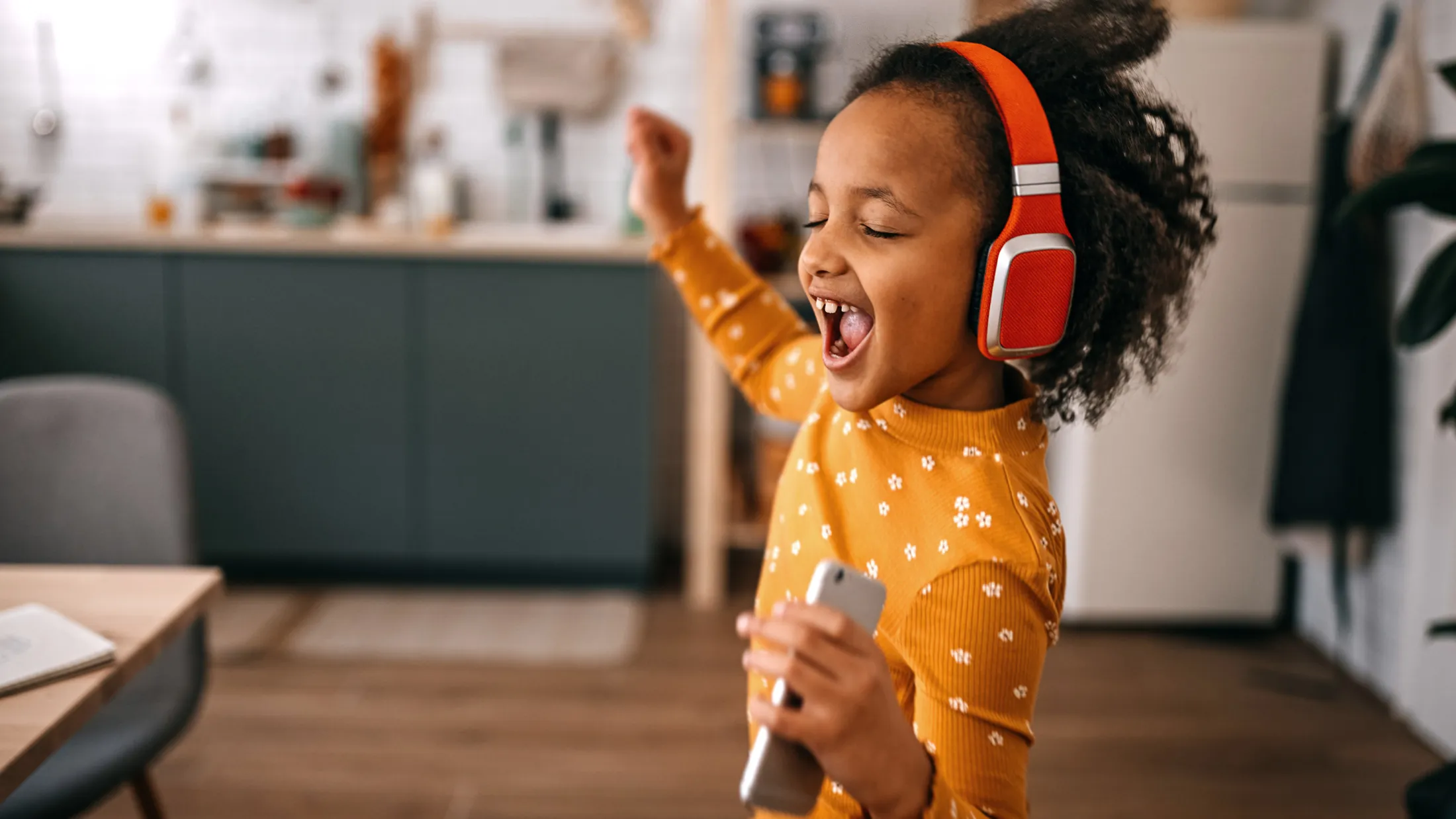 Young girl wearing red headphones and dancing joyfully in a modern kitchen, holding a smartphone.