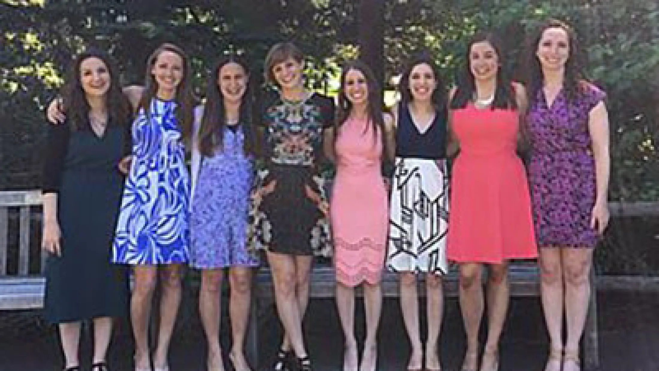 Eight women standing in a line outdoors in bright dresses, smiling in front of trees.