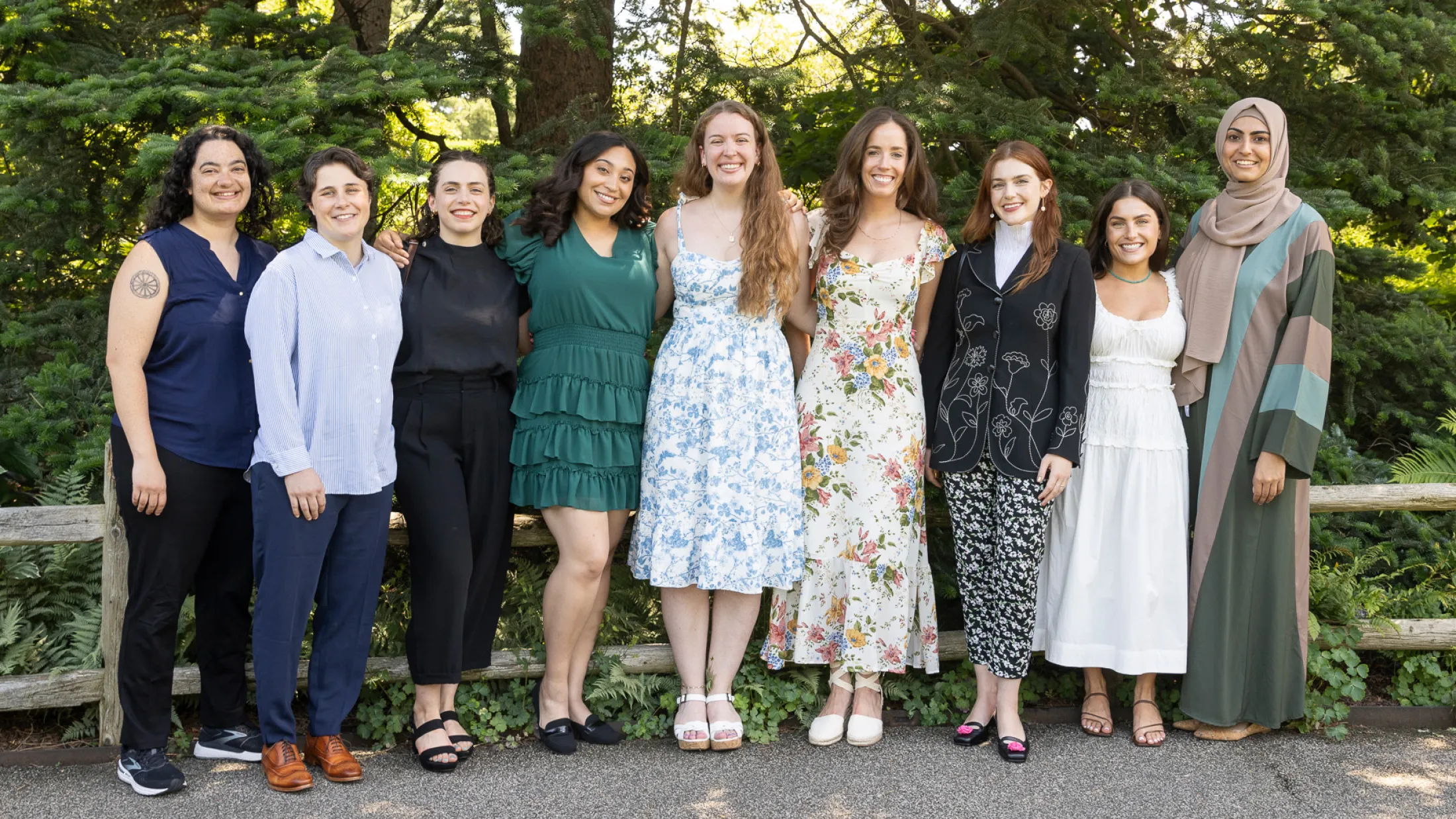 Nine individuals smiling and standing together outside on a sunny day with trees behind them.