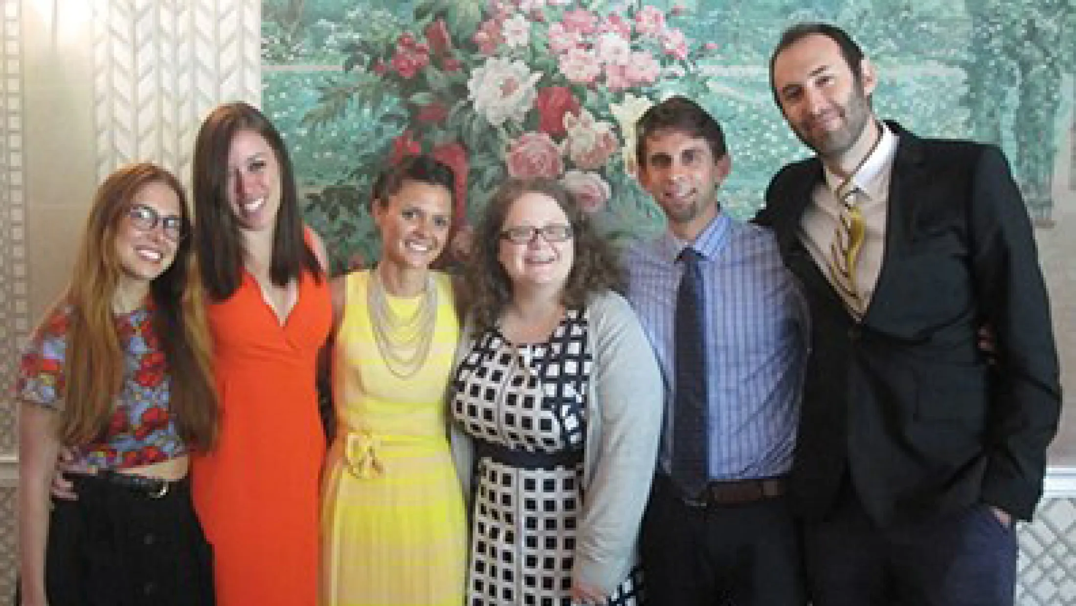 Six people standing indoors in front of a floral mural, dressed in colorful and formal clothing.