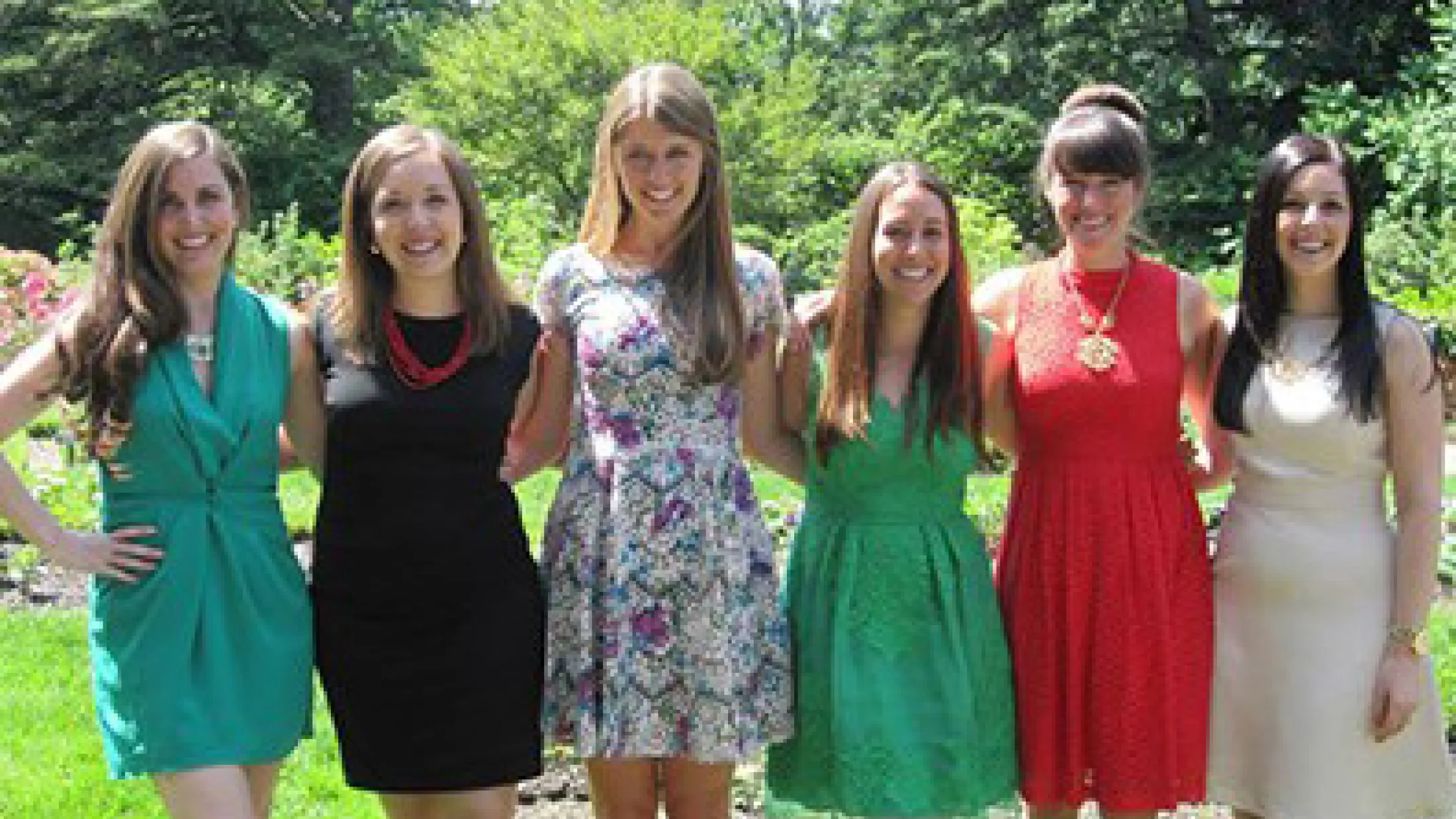Six women standing outdoors in dresses, smiling with trees and sunlight in the background.