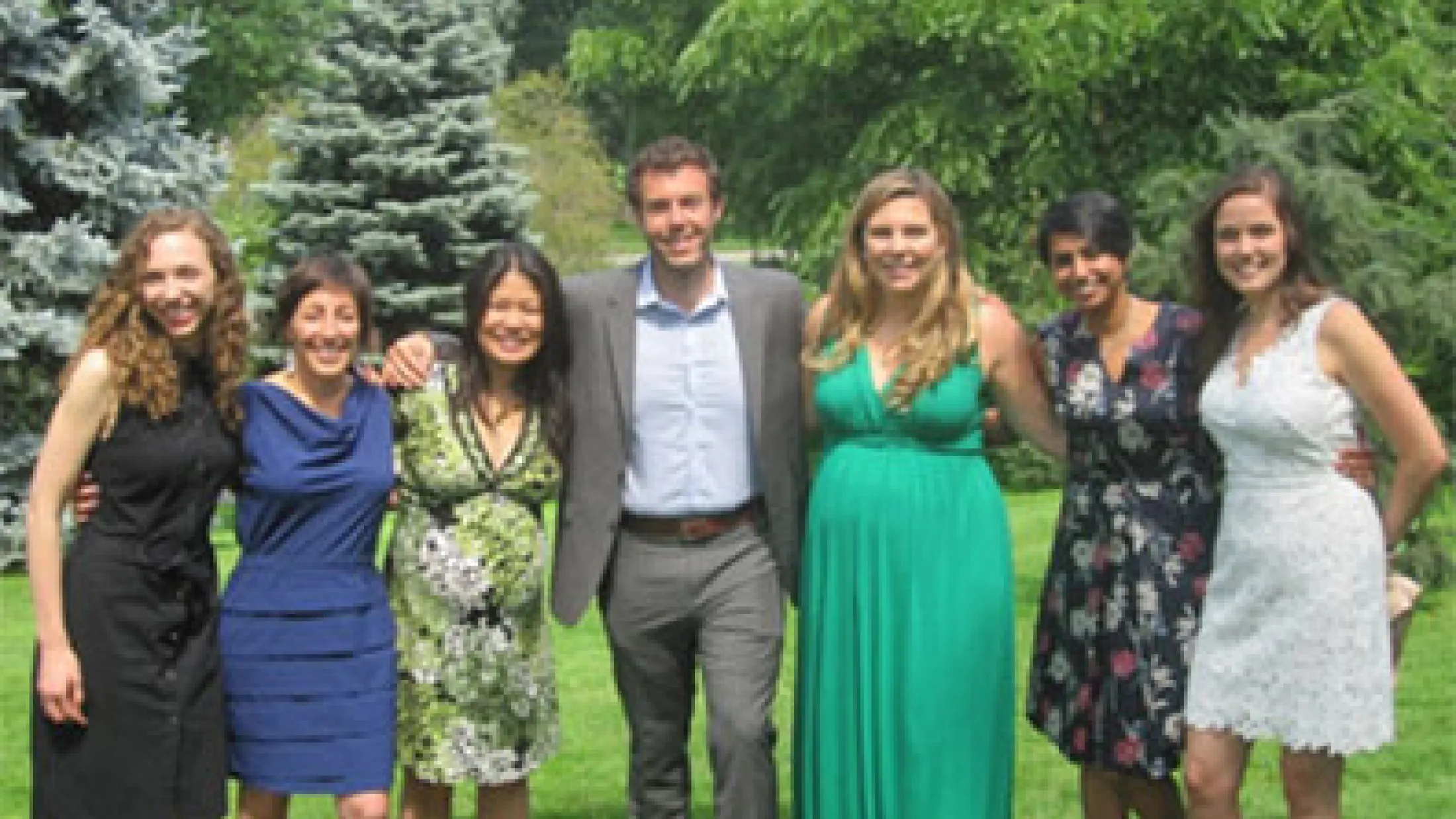 Seven people standing in a grassy area with trees behind them, smiling in formal summer outfits.