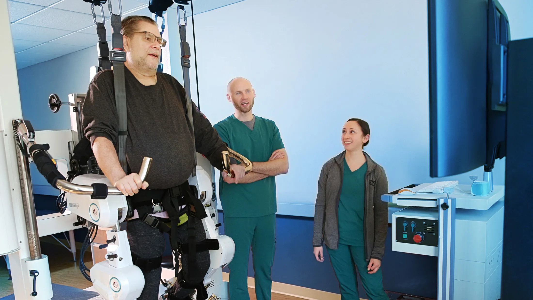 Man using a robotic gait training treadmill while two clinicians observe and guide the therapy session.