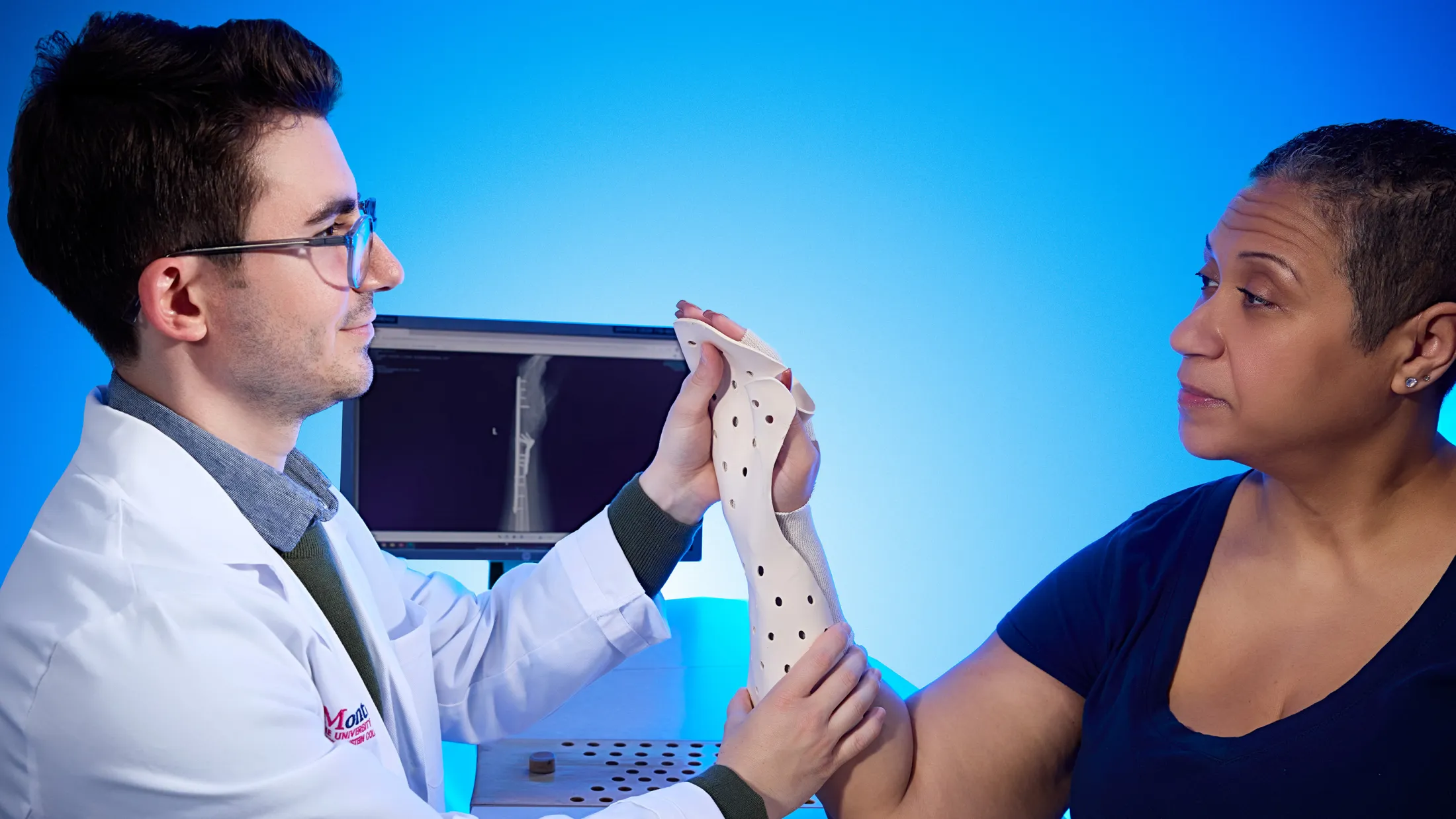 Doctor fitting a custom hand and wrist orthosis on a patient while reviewing an X-ray in the background.