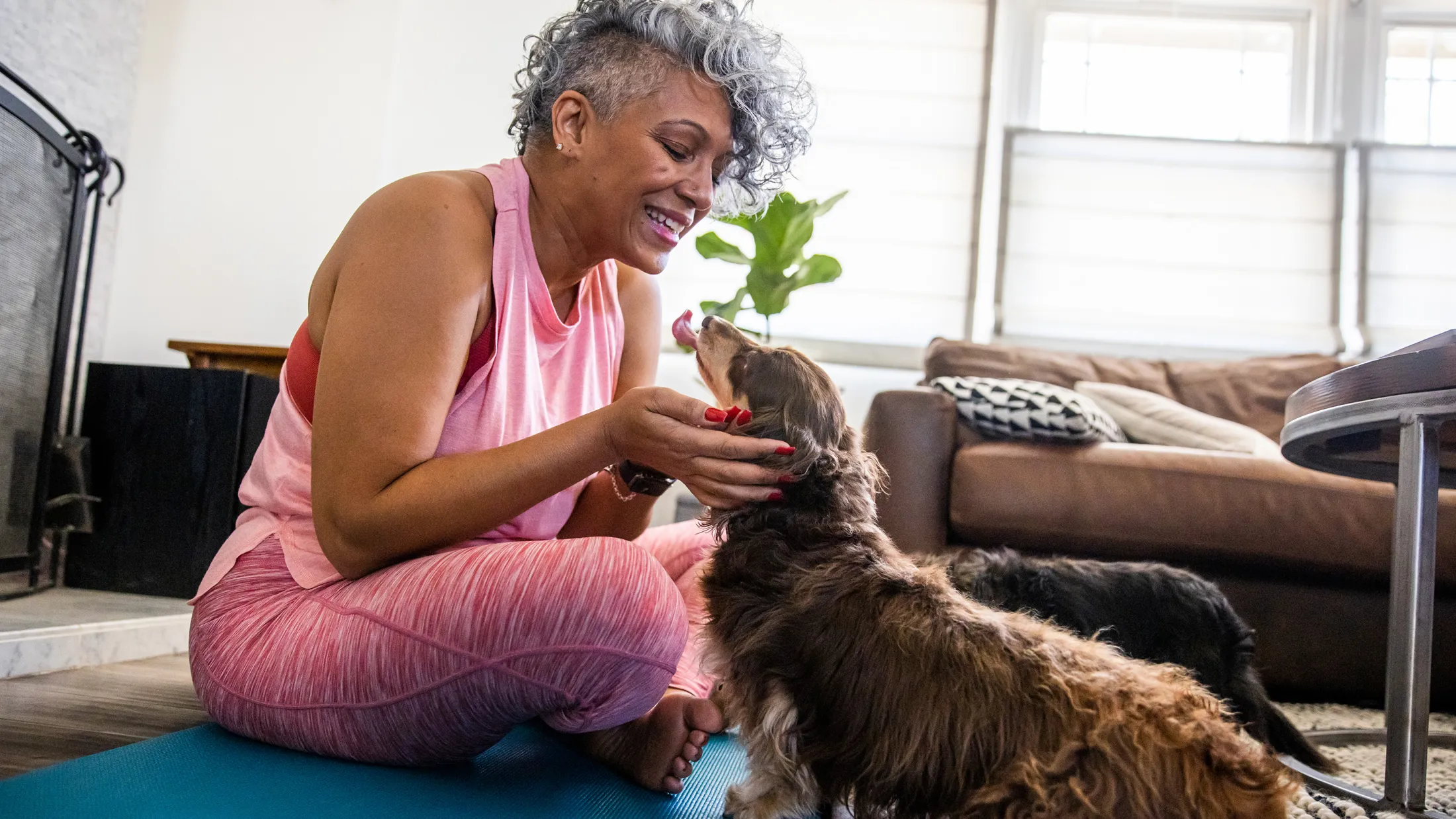 Woman in pink workout clothes smiling and playing with her dog while sitting on a yoga mat at home.