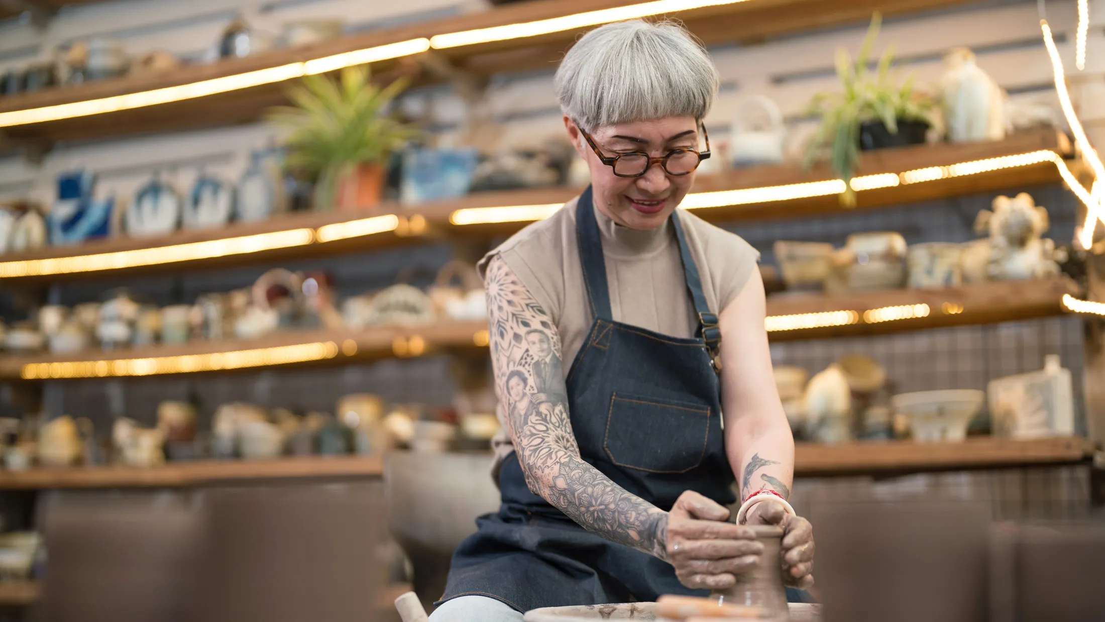 Smiling woman with gray hair and tattoos shapes clay on a pottery wheel in a ceramics studio filled with handmade pieces.