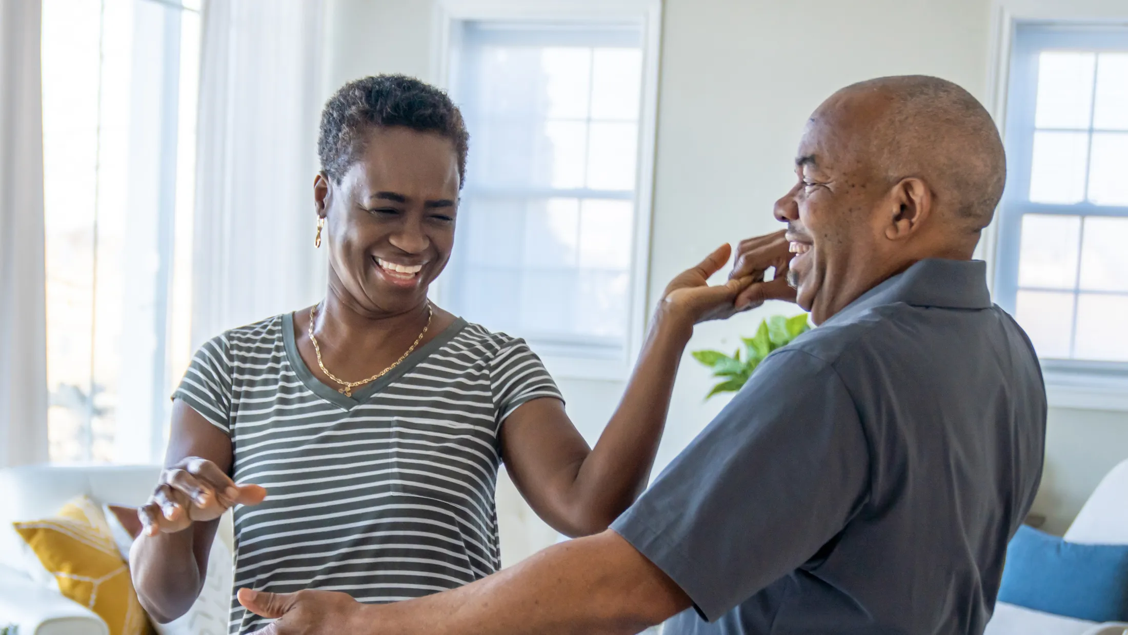 Smiling older couple dancing together in a bright, sunlit living room.