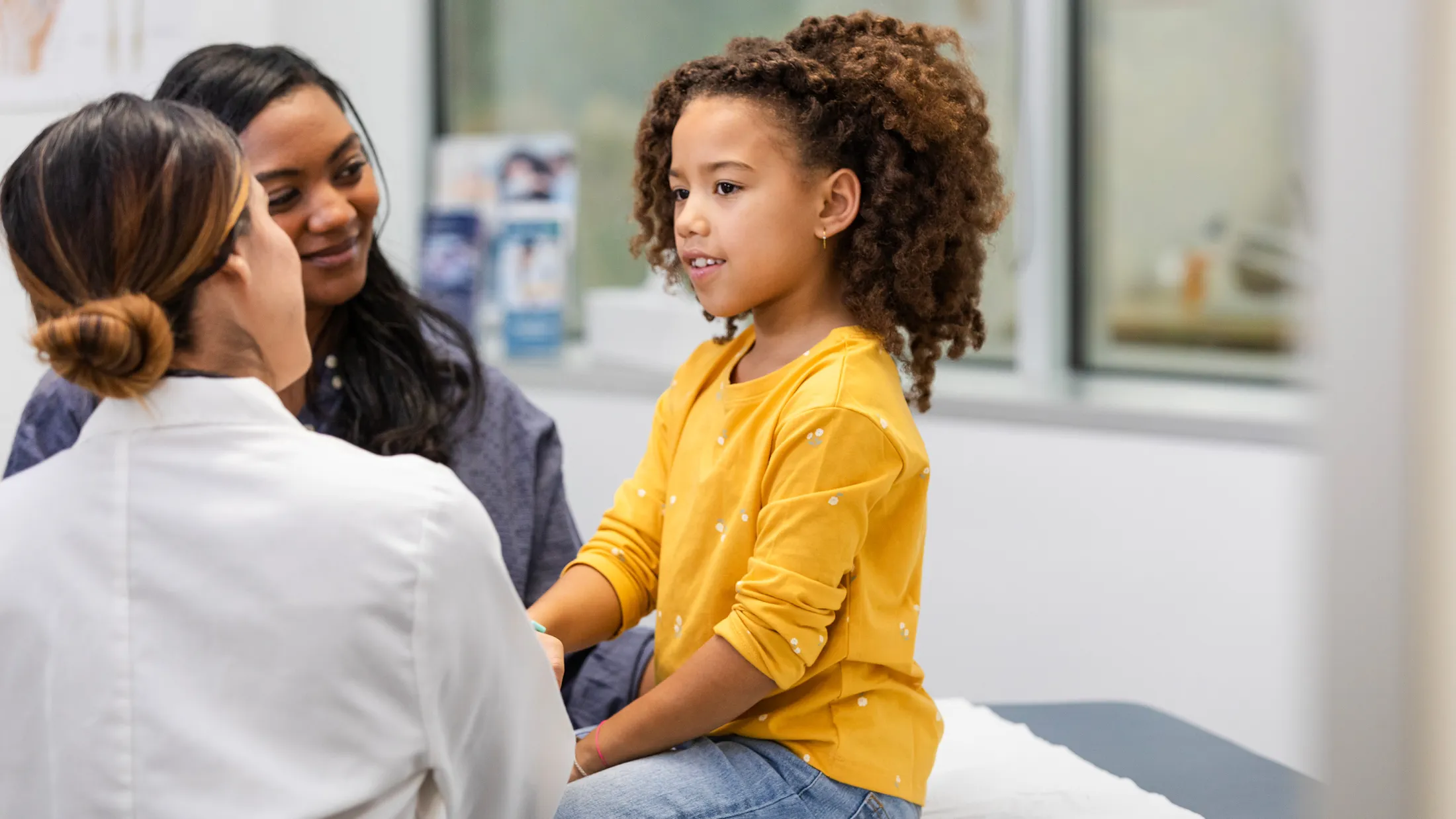 Young girl in a yellow shirt sitting on an exam table, speaking with a female doctor while her mother watches supportively.