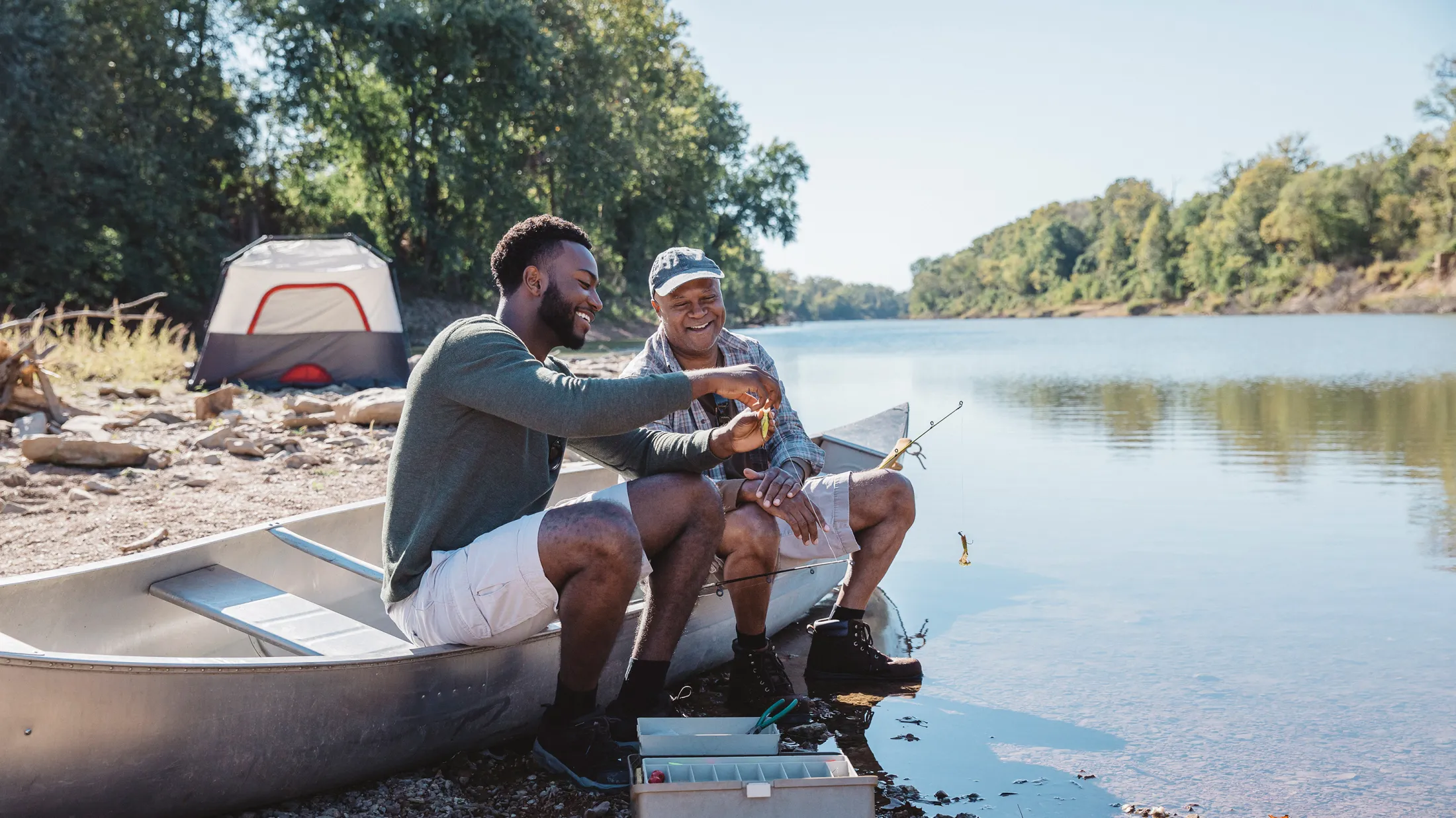 Two men smiling and preparing fishing gear while sitting in a canoe on a peaceful riverbank with a tent in the background.