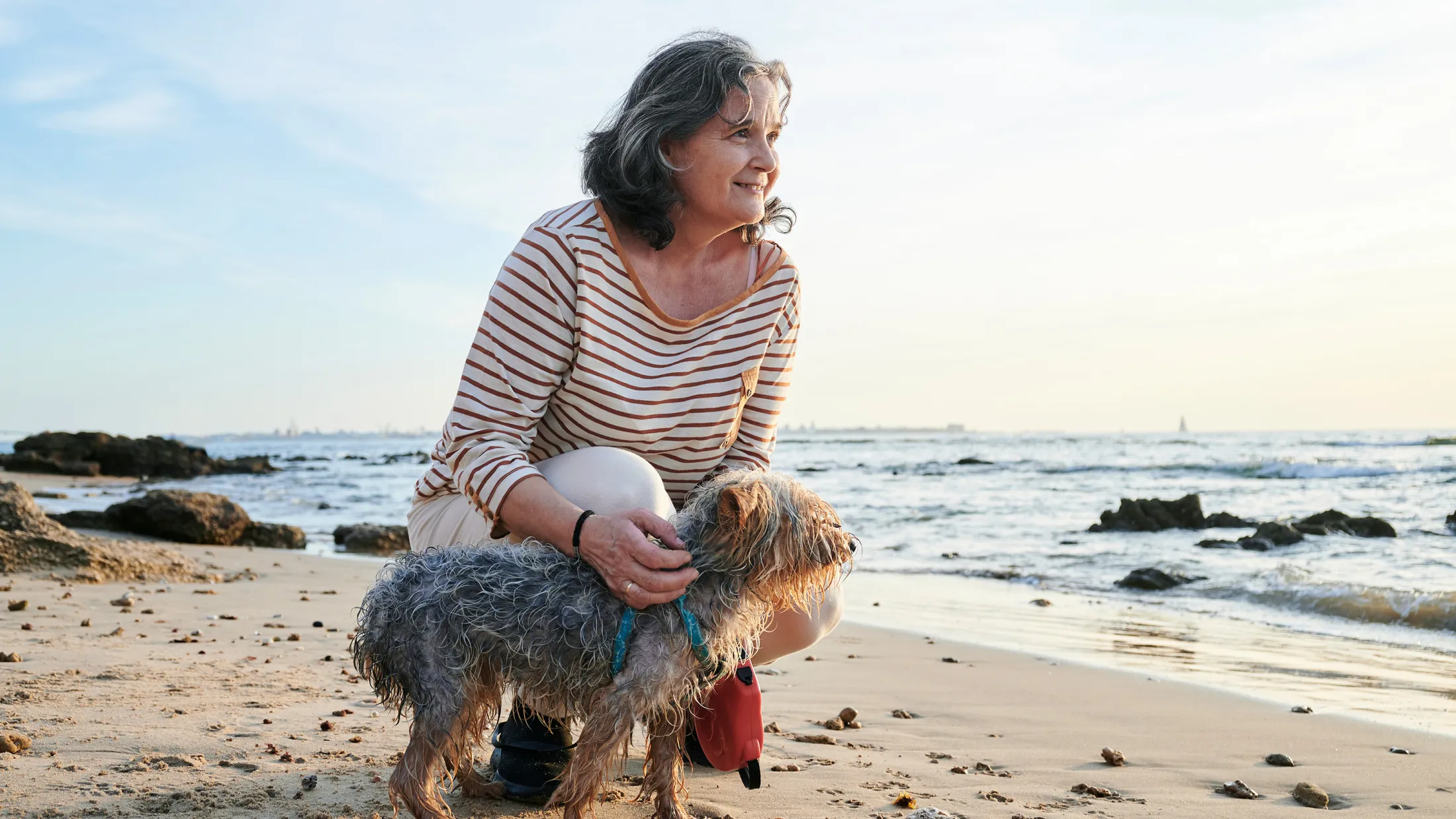 Older woman kneeling on a sandy beach next to a small, wet dog, both looking out at the ocean with a peaceful expression.