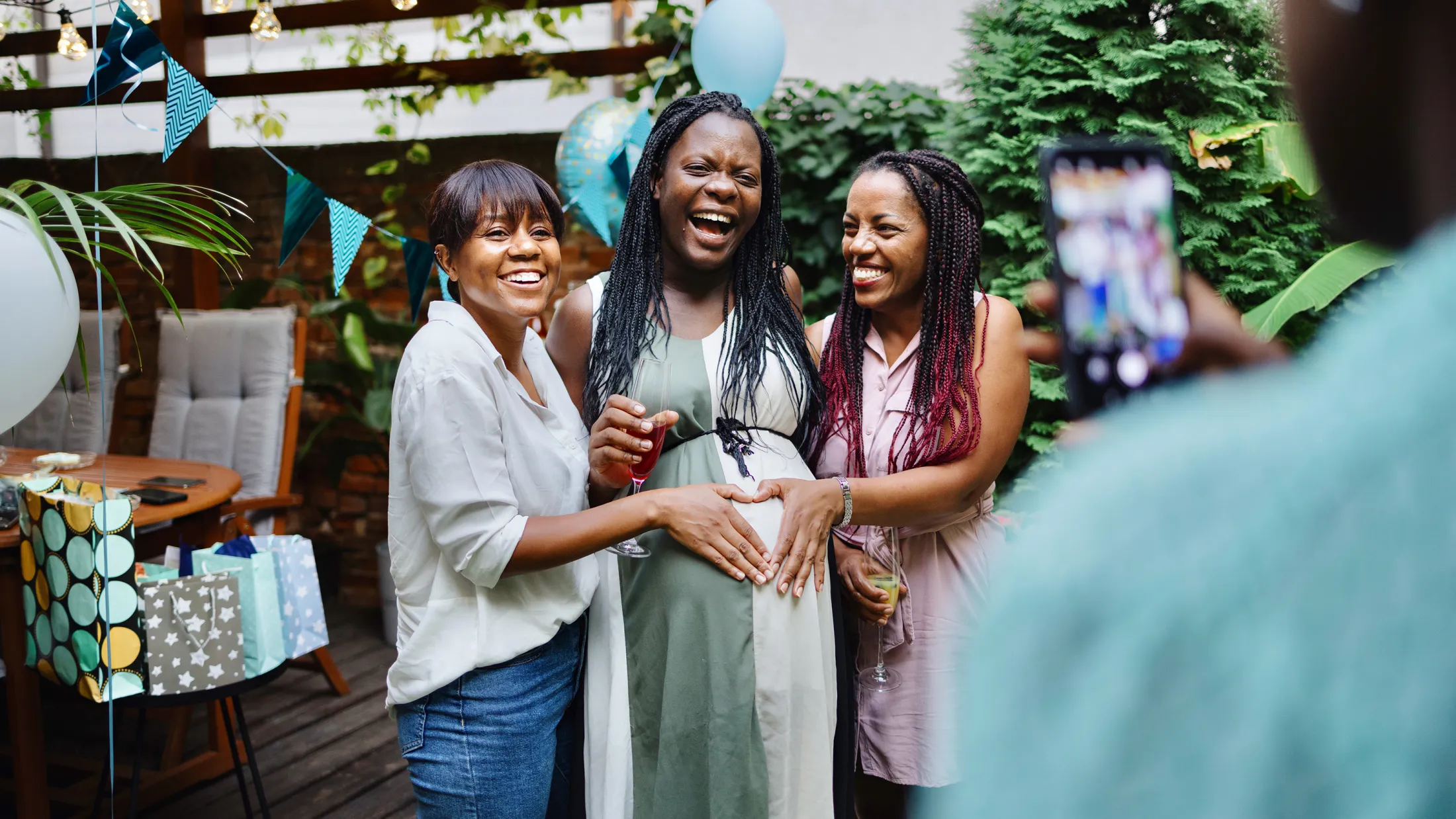 Three women at an outdoor baby shower laughing together, with one visibly pregnant woman holding a drink while the others place their hands on her belly.