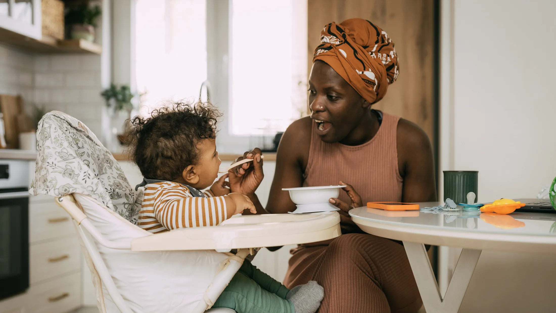 Mother sitting at a kitchen table feeding her baby in a high chair while smiling and interacting playfully.