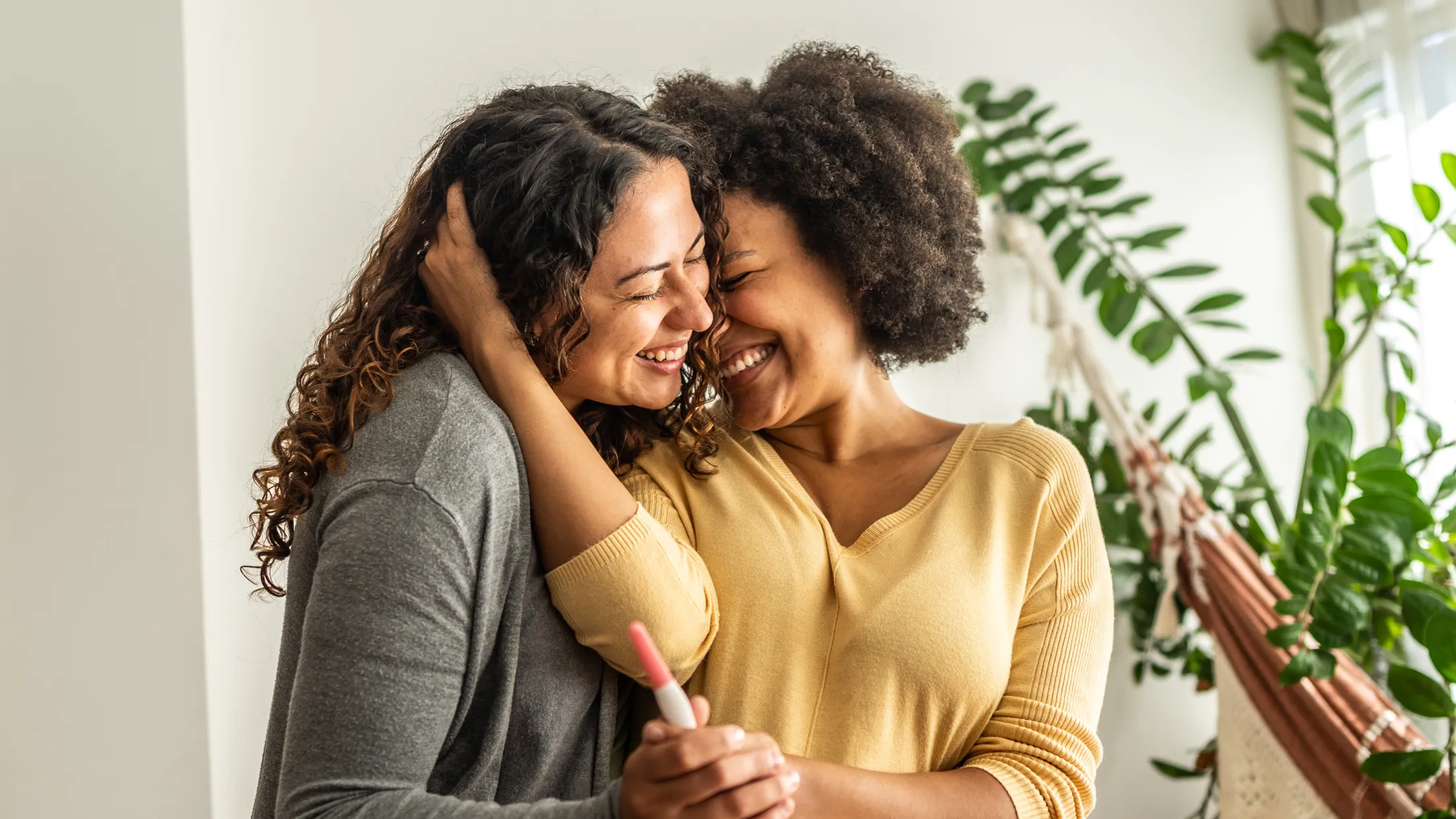 Two women embracing joyfully while holding a positive pregnancy test in a cozy, plant-filled living room.