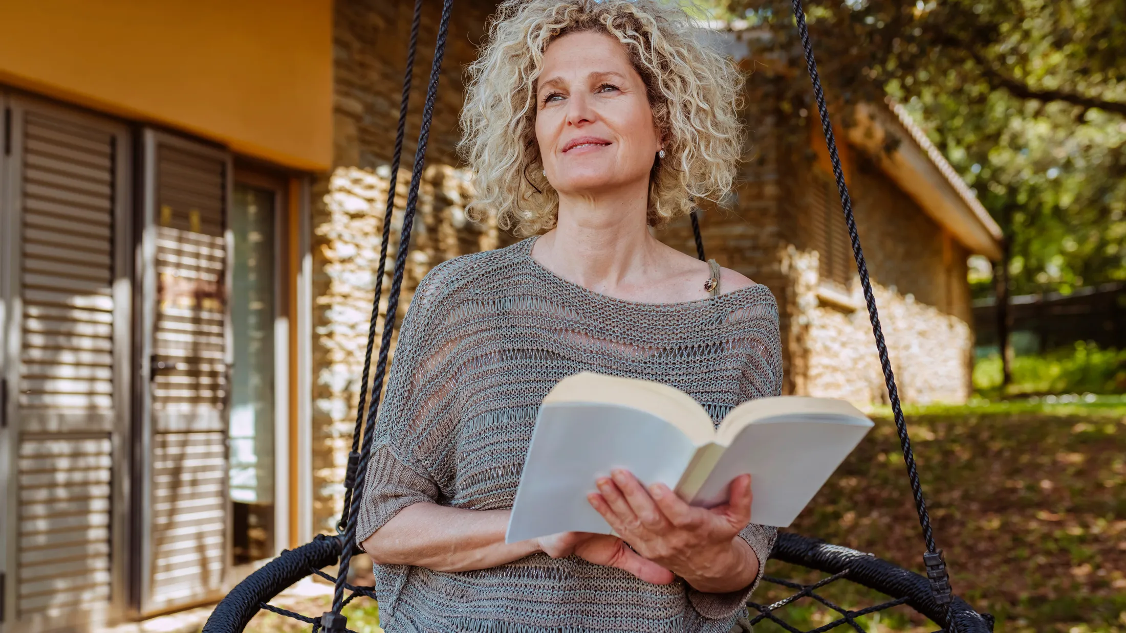 Mature woman reading a book on a hanging chair outside her home, surrounded by greenery