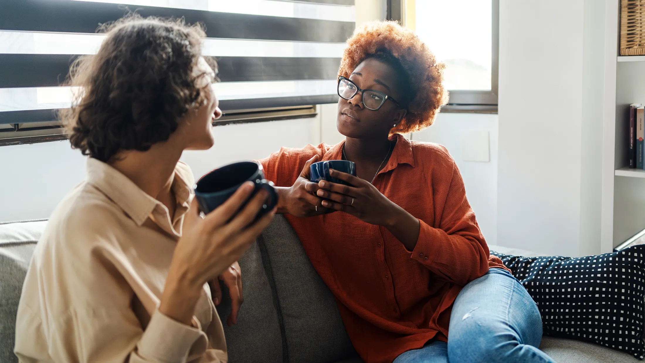 Two women sit on a couch having an intimate conversation over coffee in a well-lit home environment.