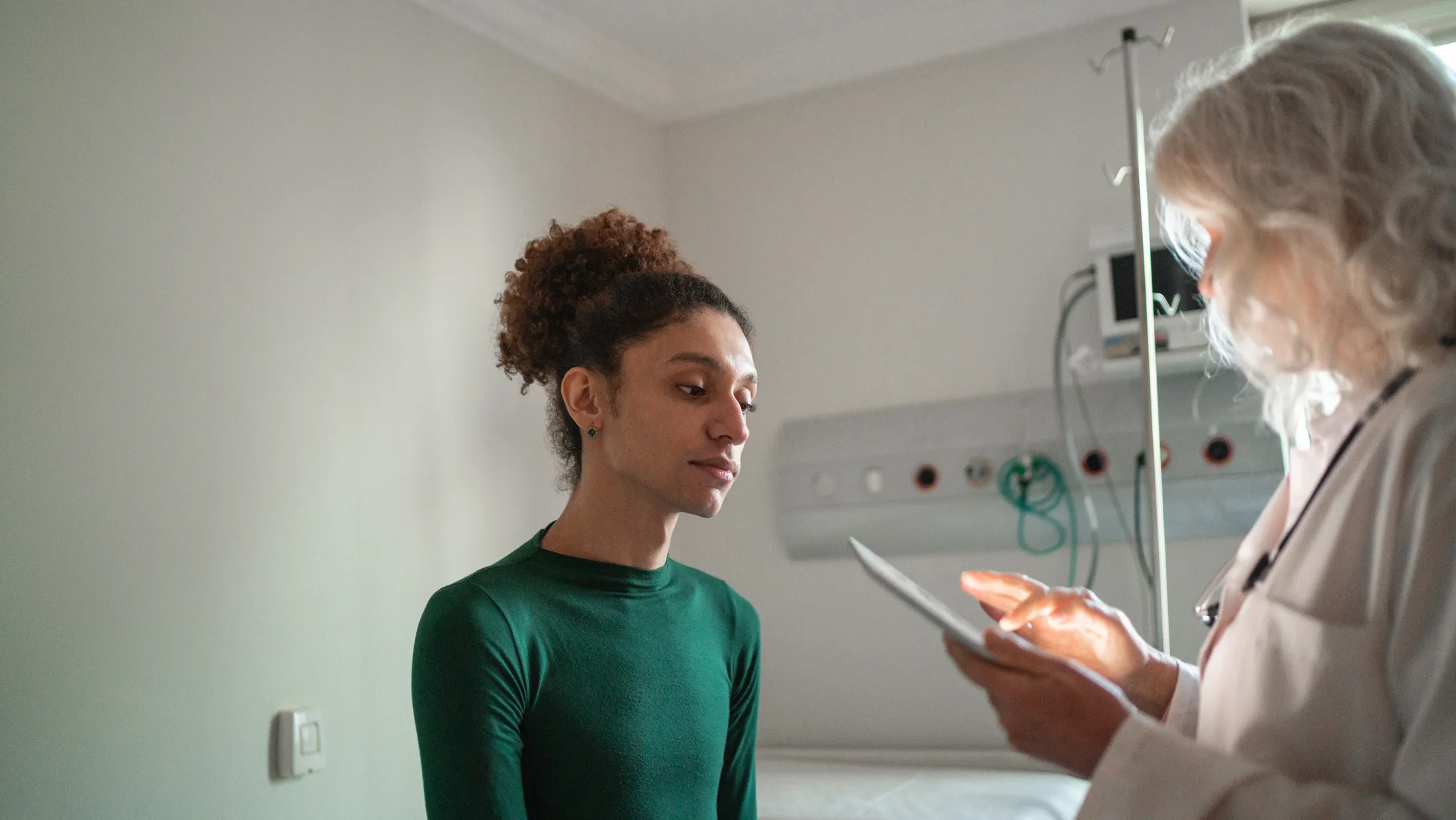 A transgender patient in a green shirt listens as a doctor reviews information on a tablet in a clinic.