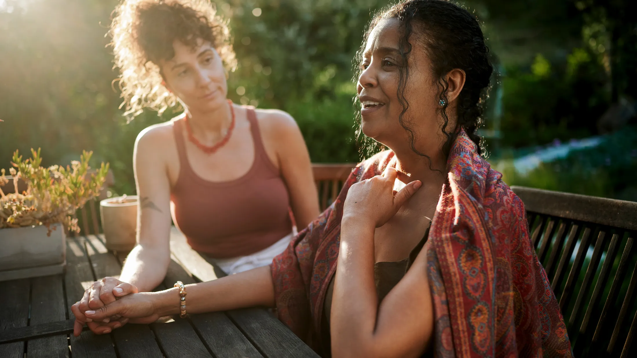 Two women sit outside at a wooden table in a sunlit garden, one woman speaking passionately with her hand on her chest while the other listens supportively, holding her hand.