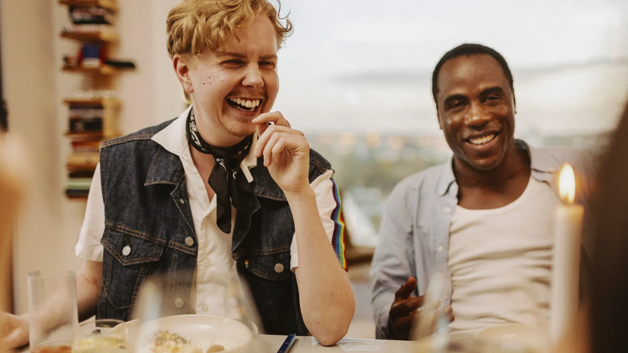 A transgender man laughing with friends over a shared meal, enjoying conversation at a dinner table.