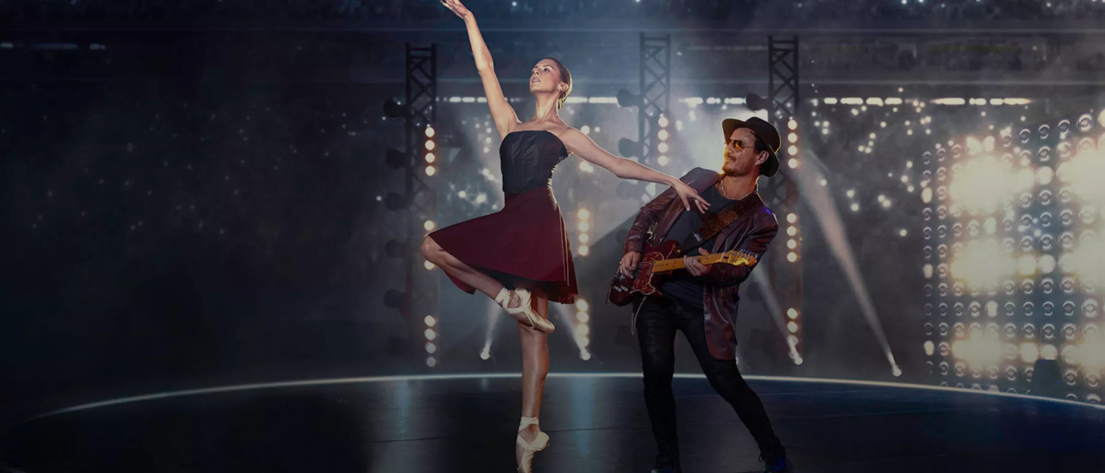 Chiara dances ballet in a deep burgundy skirted ballet dress with black bodice, next to a man playing an electric guitar. All around them, lights sparkle.