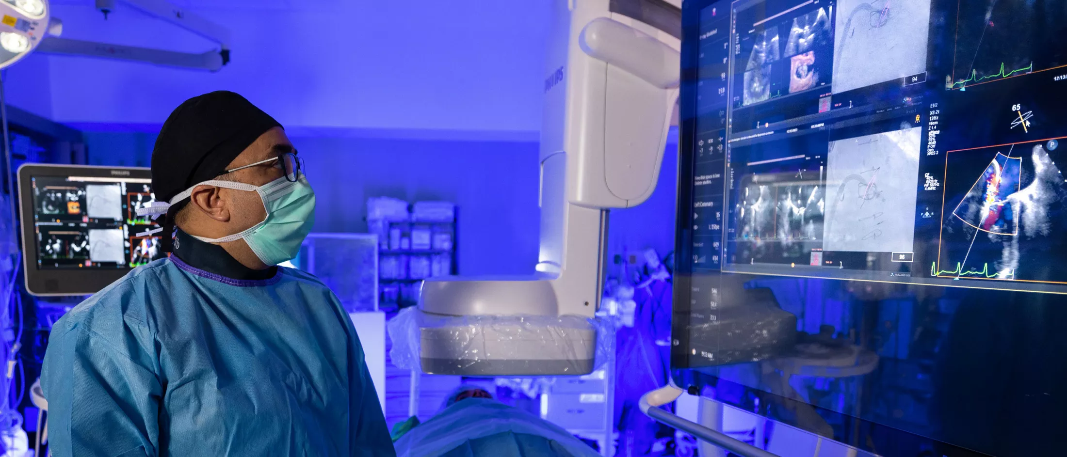 A medical professional in scrubs and a mask observes diagnostic images on multiple monitors in a high-tech medical facility.