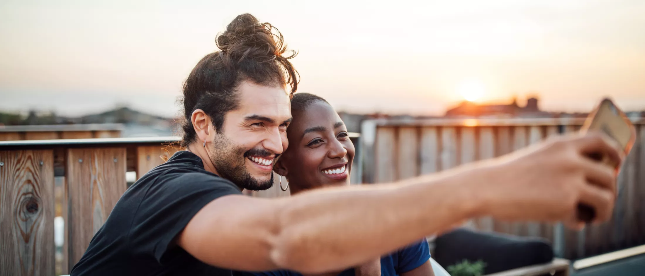 A white man with a beard and long hair in a bun poses for a selfie with a short-haired black woman near a wooden bridge.