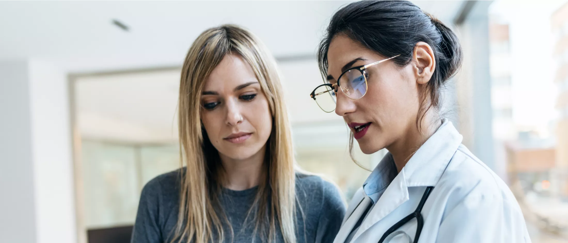 A white woman with long blonde hair in a dark blue shirt looks at an offscreen tablet while listening to a female doctor.