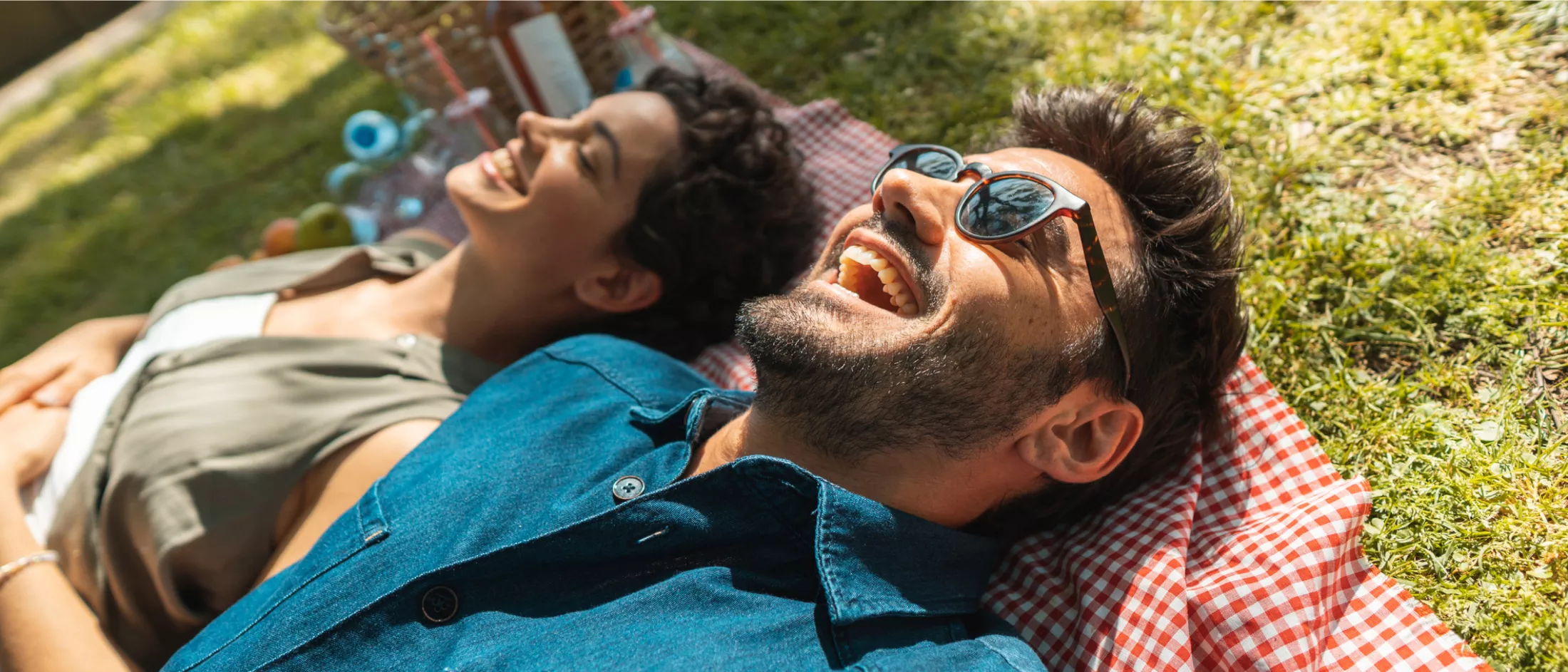 A young couple lay on a gingham picnic blanket with a picnic basket and food between them.